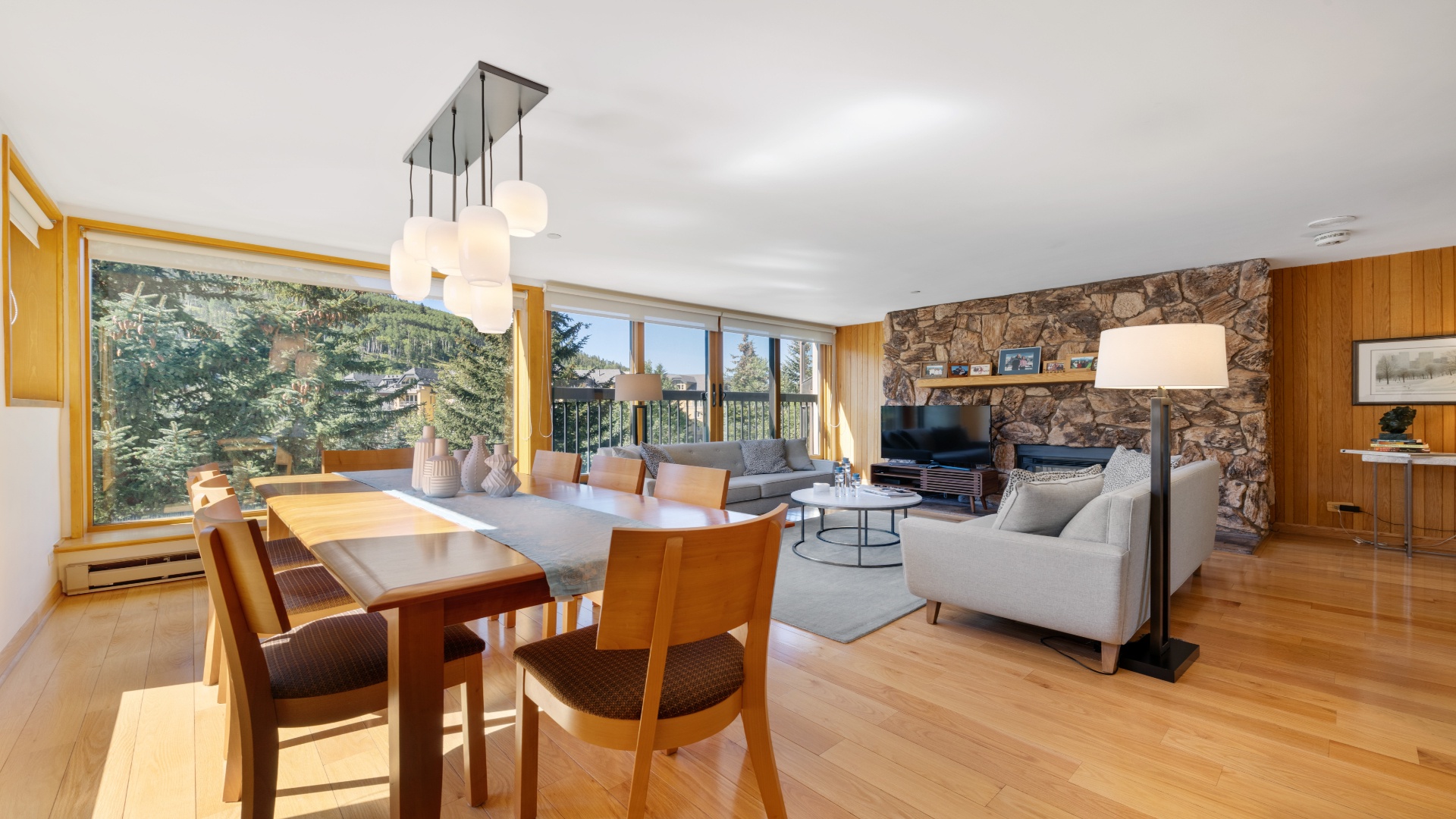 Sunlight pours through floor-to-ceiling windows into your open-plan living space, where a cozy stone fireplace, modern furnishings, and warm wood accents create the perfect mountain retreat.