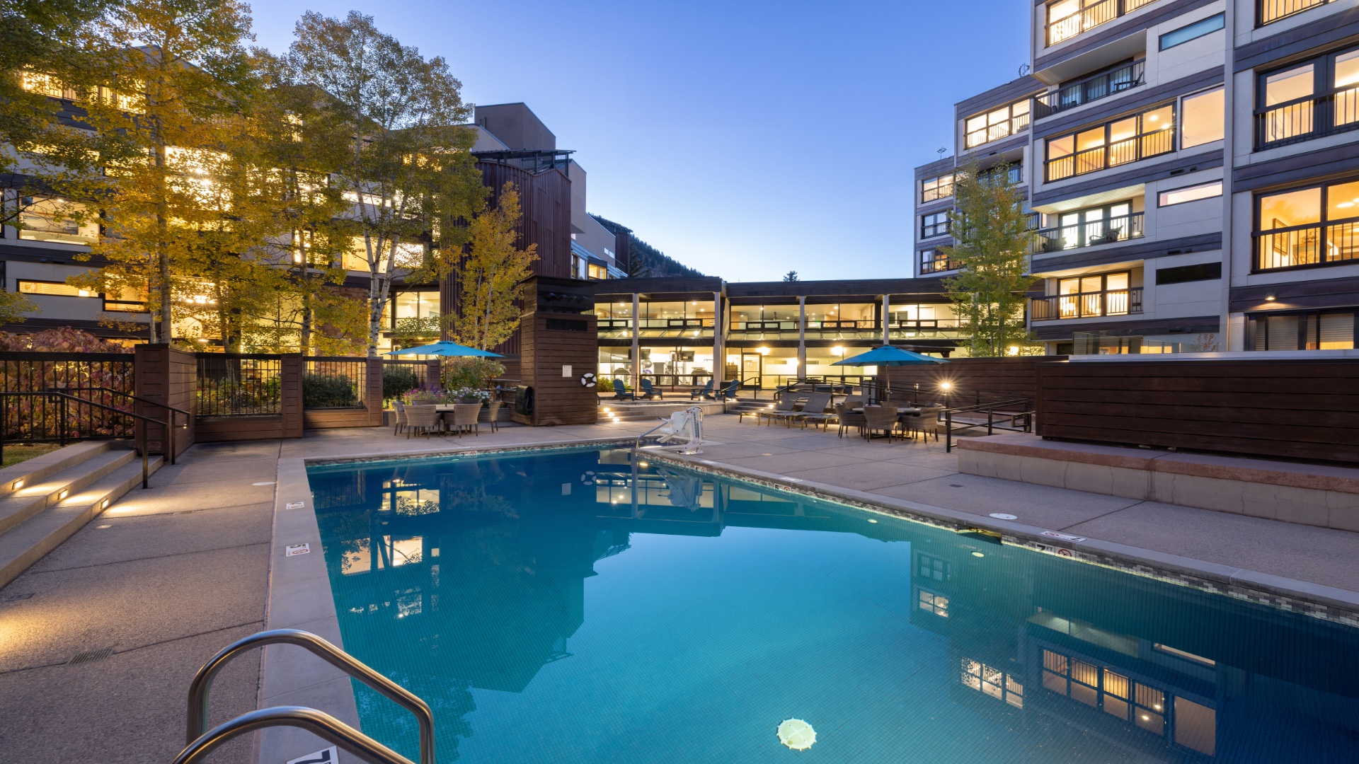 A modern apartment complex surrounds a pristine outdoor pool, with contemporary architecture, landscaped grounds, and ambient evening lighting creating an inviting urban retreat.