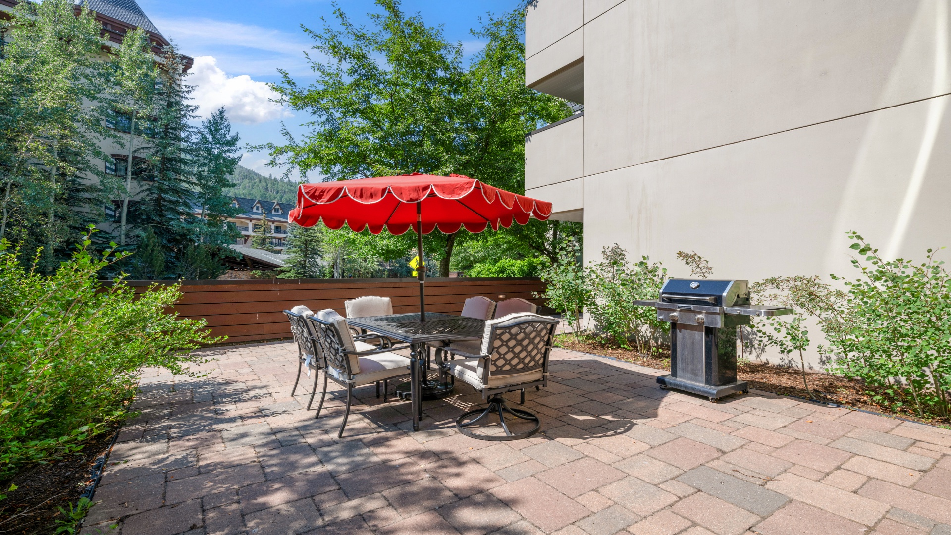 Your private patio invites mountain-fresh air and sunshine while you dine alfresco at the fully-equipped outdoor table, shaded by a vibrant red umbrella, with a modern grill ready for your cookout.
