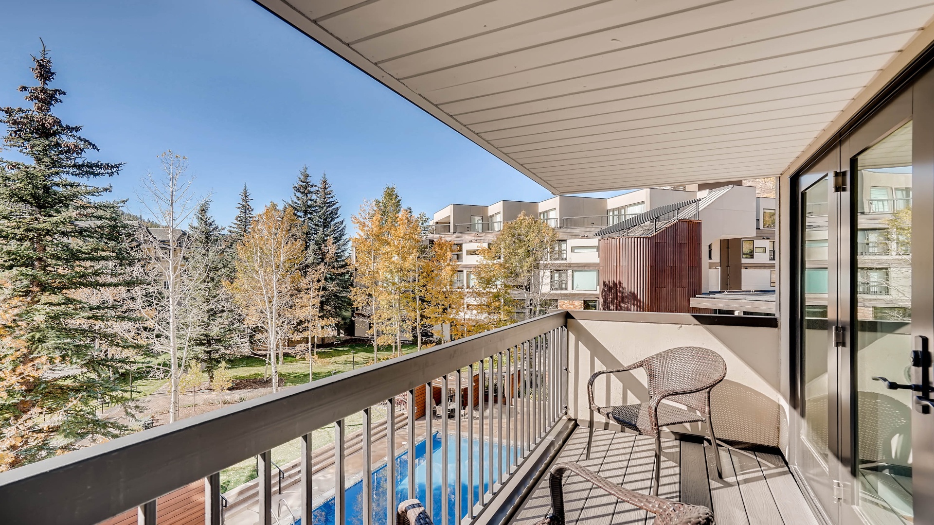 A covered balcony overlooking a peaceful residential neighborhood surrounded by evergreens and trees showing autumn color, with nearby mid-rise buildings and a pool area visible below.