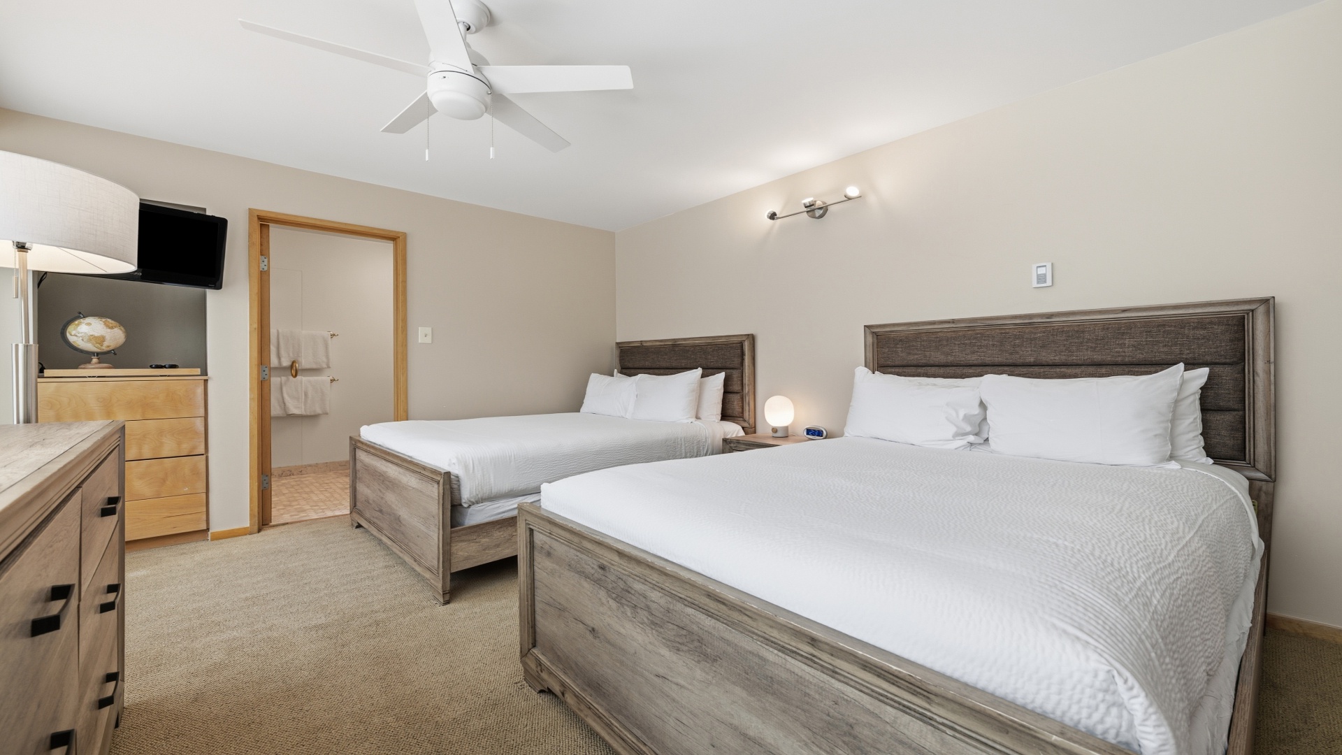 Your spacious bedroom features two plush beds with fresh white linens, a ceiling fan for comfort, and a mounted TV for your entertainment. Warm lighting and neutral tones create a peaceful retreat after a day of adventure.