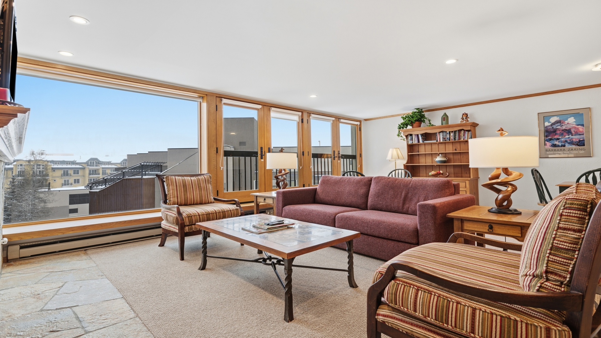 Relax in your sunlit living room with panoramic windows framing city views. Comfortable seating, warm wood accents, and plenty of natural light create a welcoming space.