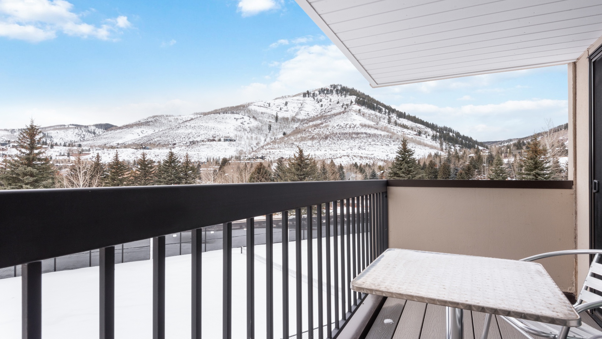 A snowy mountain view with forested slopes and evergreens frames the property, showcasing a pristine winter landscape. The covered balcony provides a quiet vantage point to admire this tranquil alpine setting.