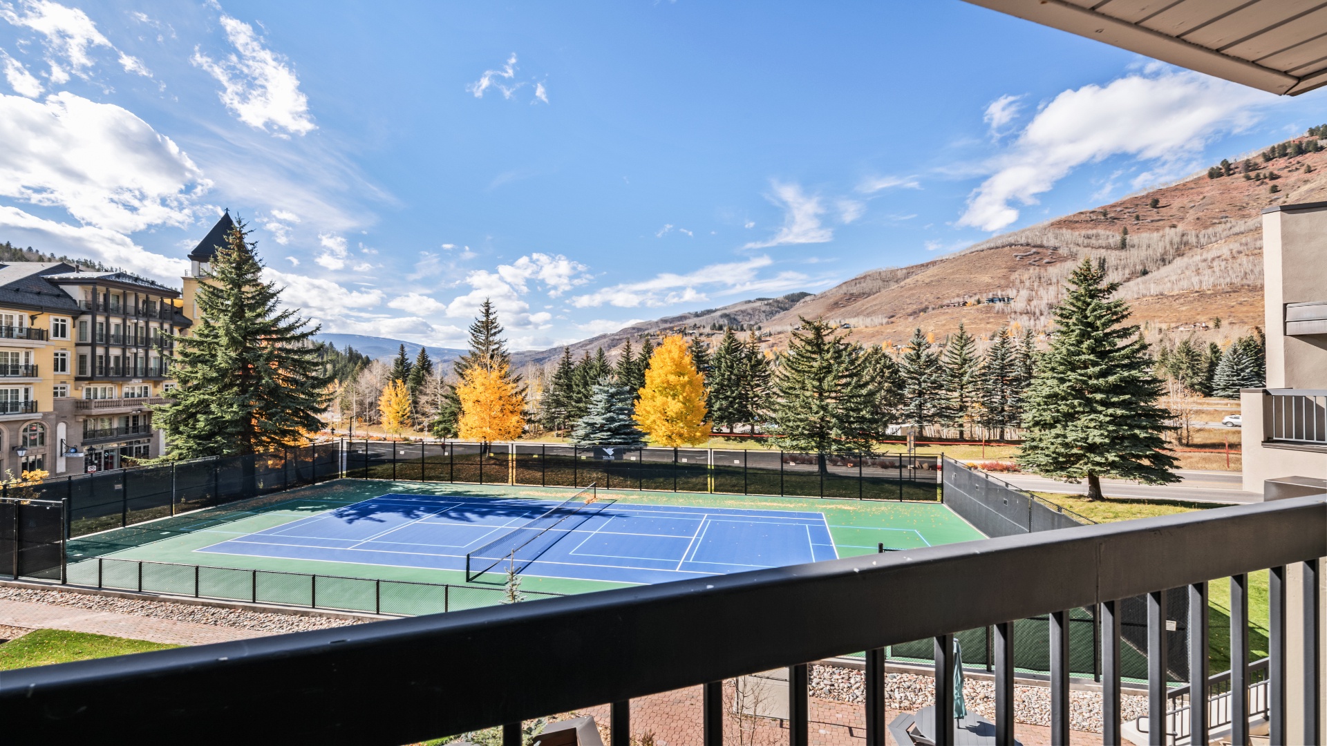 Private balcony view overlooking tennis court, evergreens, and mountain slopes beneath blue skies—ideal for morning coffee while watching a friendly match or simply soaking in the surrounding beauty.