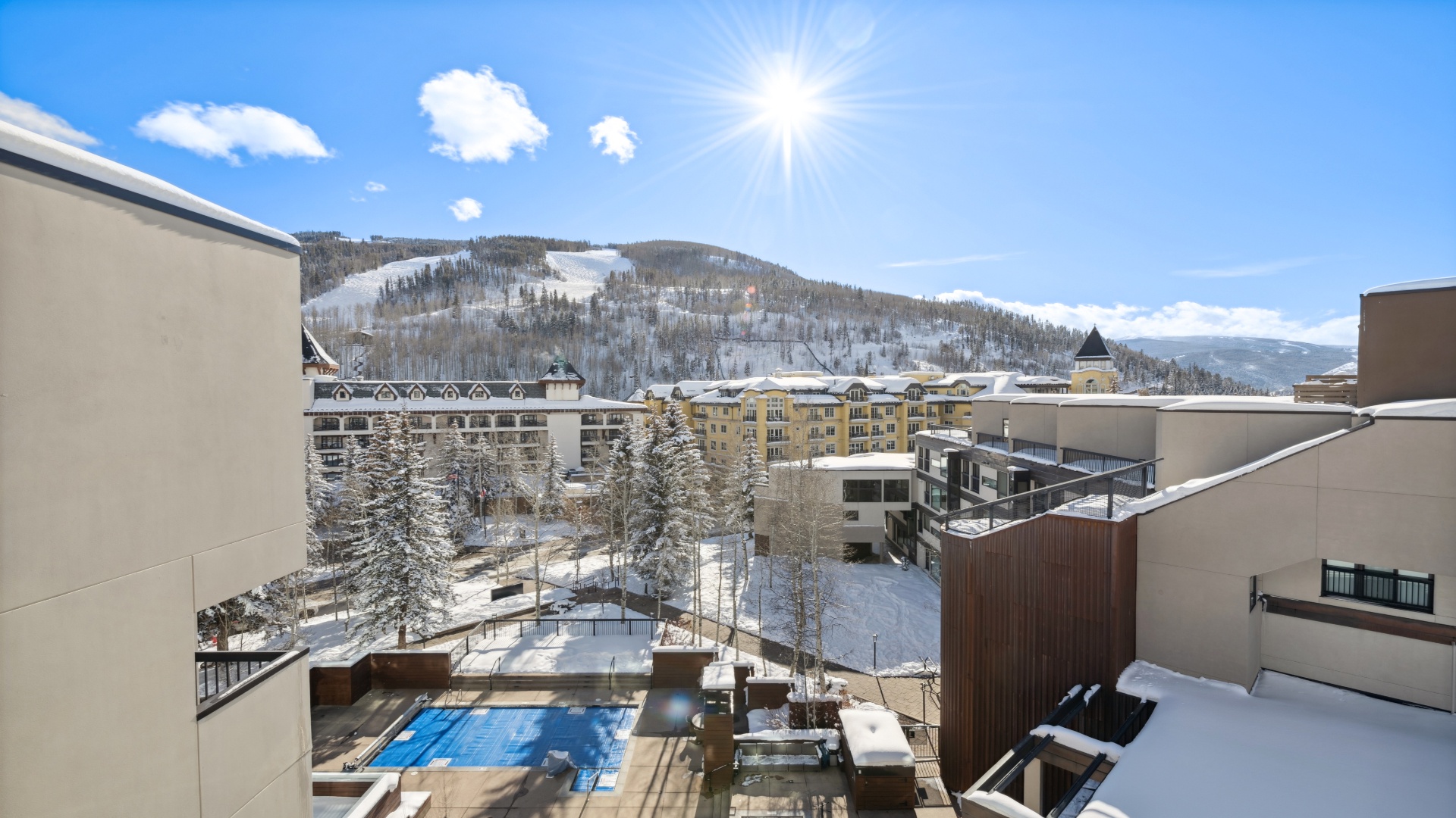 Mountain resort nestled in a winter wonderland with snow-covered slopes, a sparkling outdoor pool, and evergreen trees framing the alpine village below under bright sunshine.