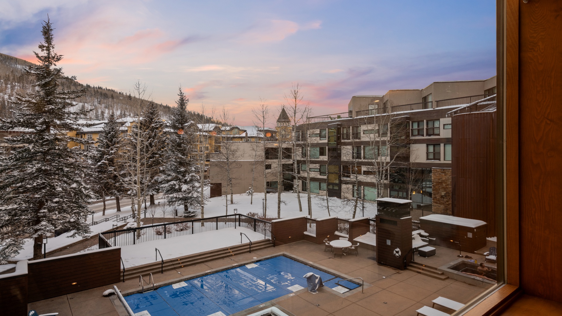 A winter mountain resort framed by evergreens and illuminated by soft sunset hues, featuring outdoor pool and hot tub facilities amid a snowy alpine setting.