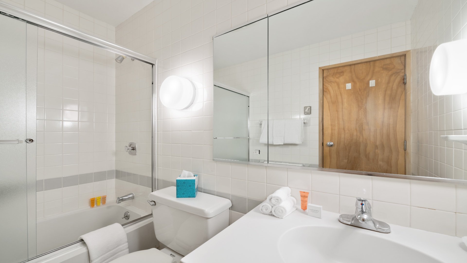 Step into your bright, spotless bathroom with a full tub-shower combo, crisp white tiles, and fresh towels waiting. The mirrored walls reflect the clean lines and modern simplicity that make you feel right at home.