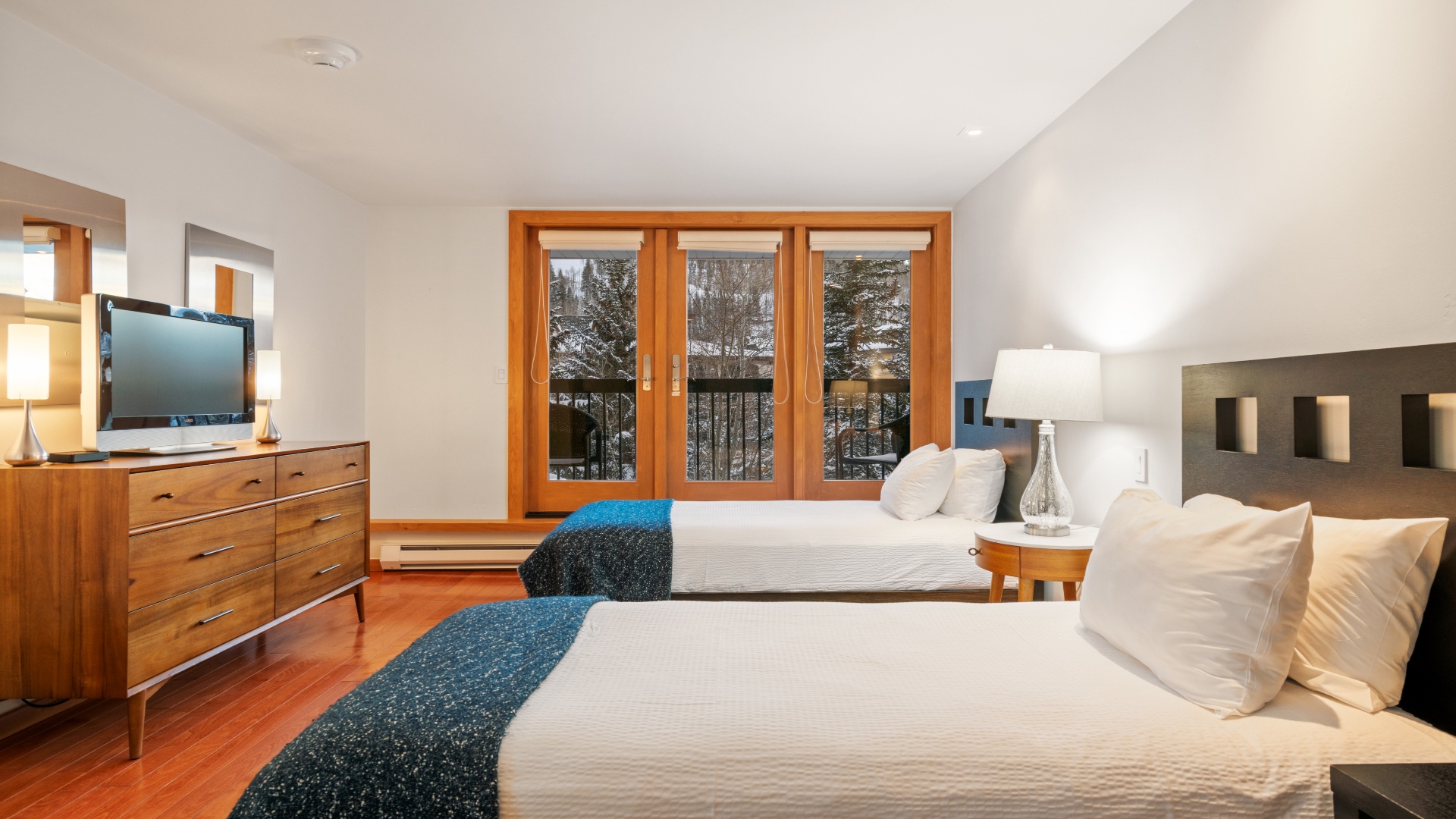 Unwind in your peaceful retreat with two plush beds, warm wood accents, and calming winter views framed by elegant balcony doors—your perfect escape awaits.