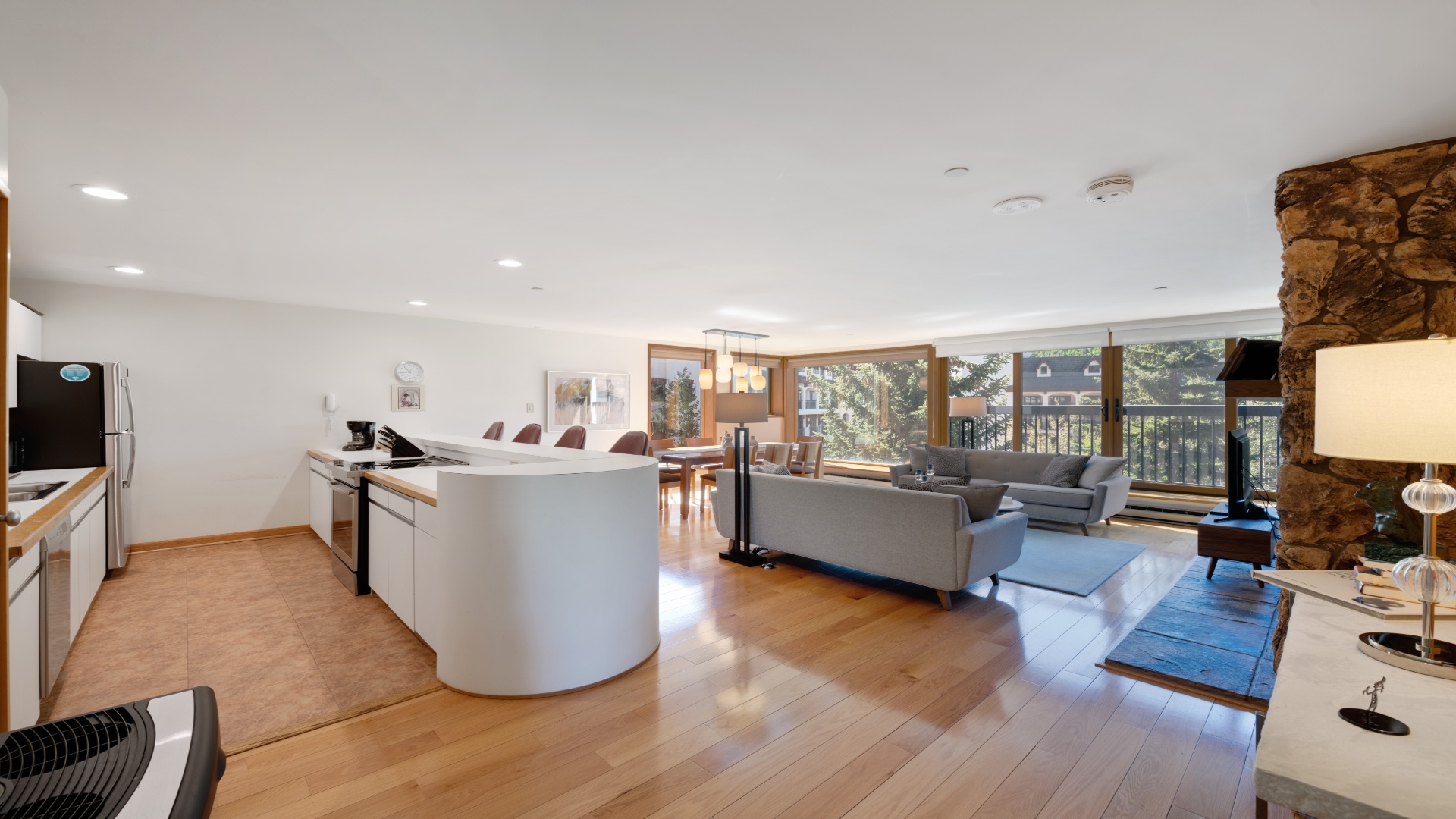 This bright, open-concept space blends kitchen, dining, and living areas, with warm wood floors, a cozy stone fireplace, and windows framing tranquil wooded views.