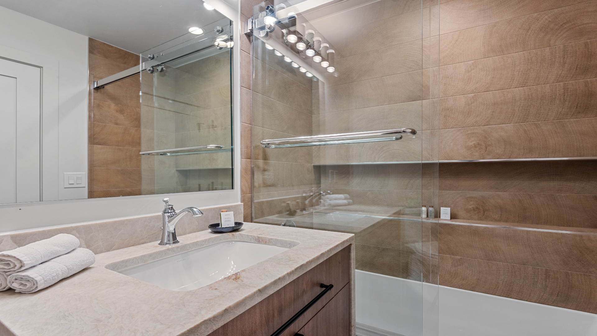 Your sleek private bathroom features warm wood-look tile, a spacious vanity, and a modern glass-enclosed tub-shower combo—ready for your relaxing soak.