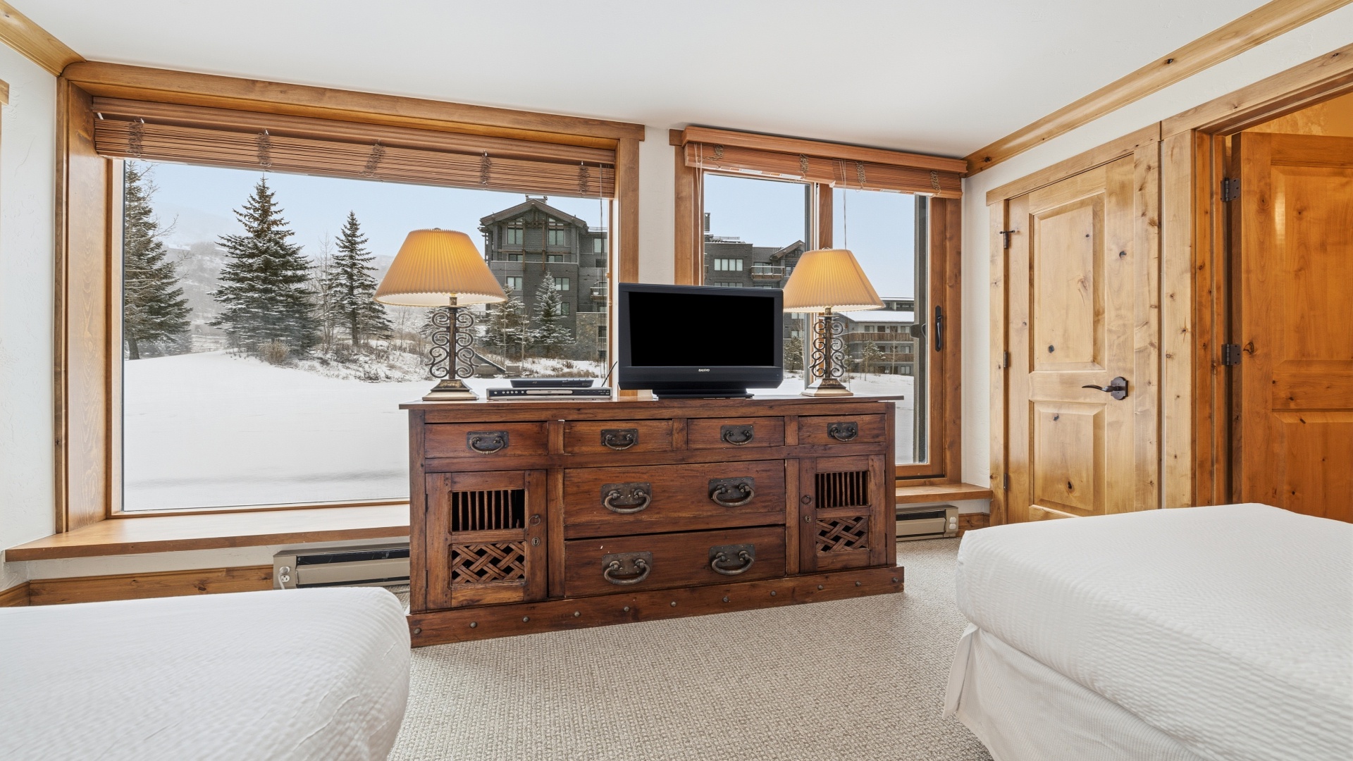 Cozy bedroom with stunning snowy mountain views through large windows, rustic wooden dresser with TV, warm lamps, and peaceful winter surroundings to enjoy during your stay.