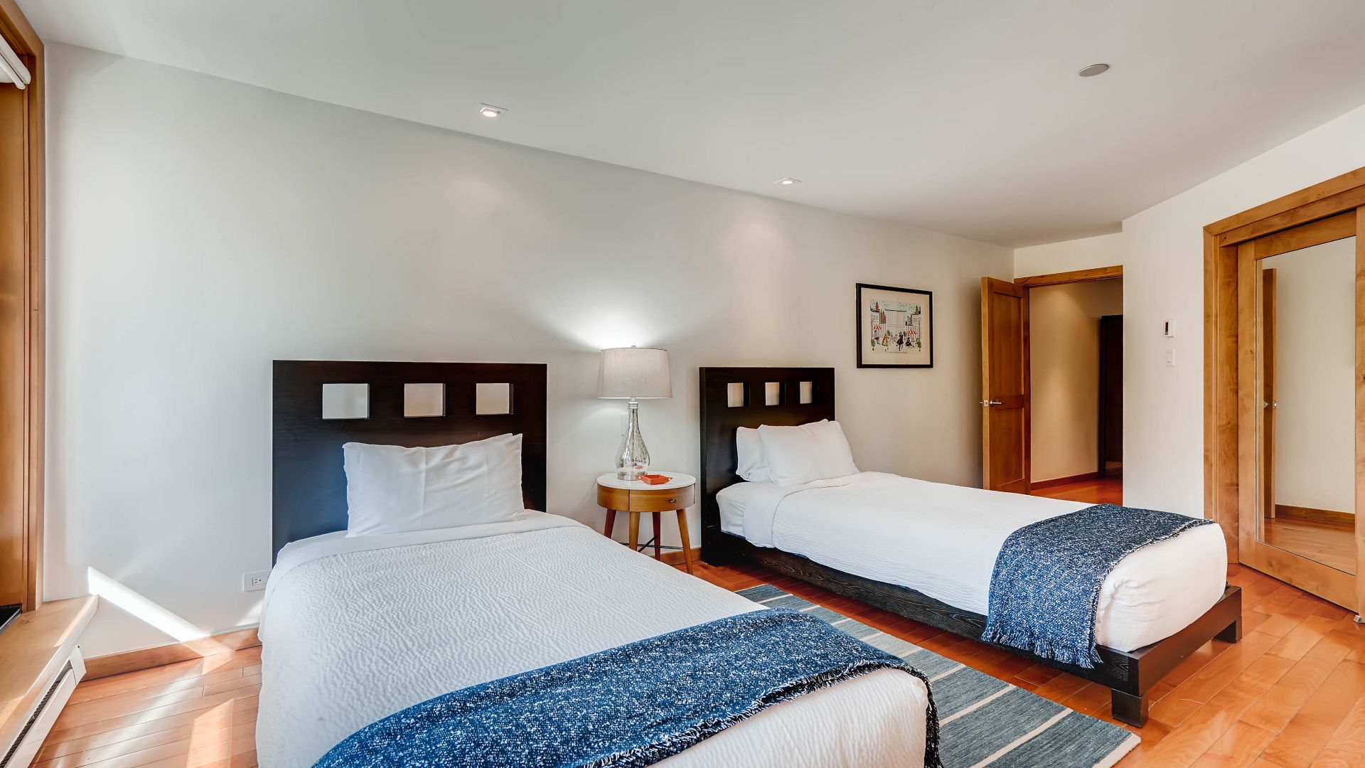 Two inviting beds with crisp white linens and cozy throws await your arrival, offering flexible sleeping arrangements in a bright, serene space with warm wood accents.