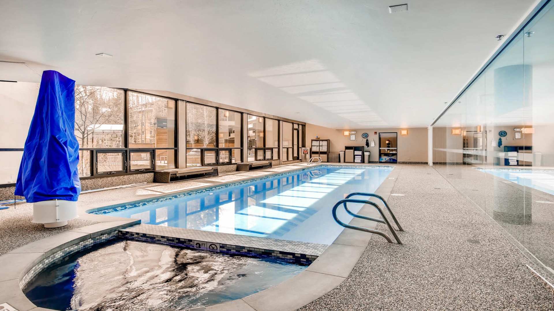 The indoor pool area offers a spacious, light-filled space to unwind, with a sleek lap pool, bubbling hot tub, and floor-to-ceiling windows overlooking the city.