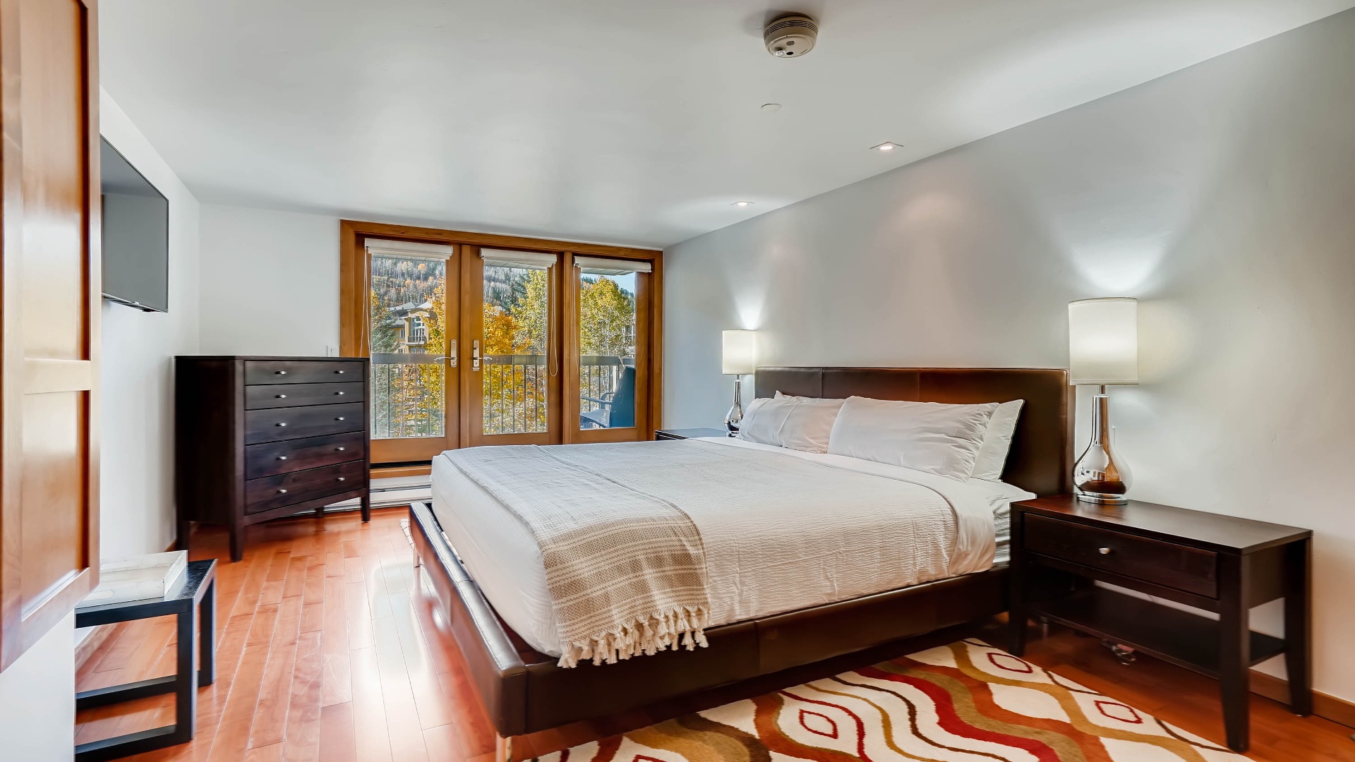 Unwind in your serene bedroom sanctuary with plush bedding, warm wood tones, and stunning autumn views through French doors that open to your private balcony.