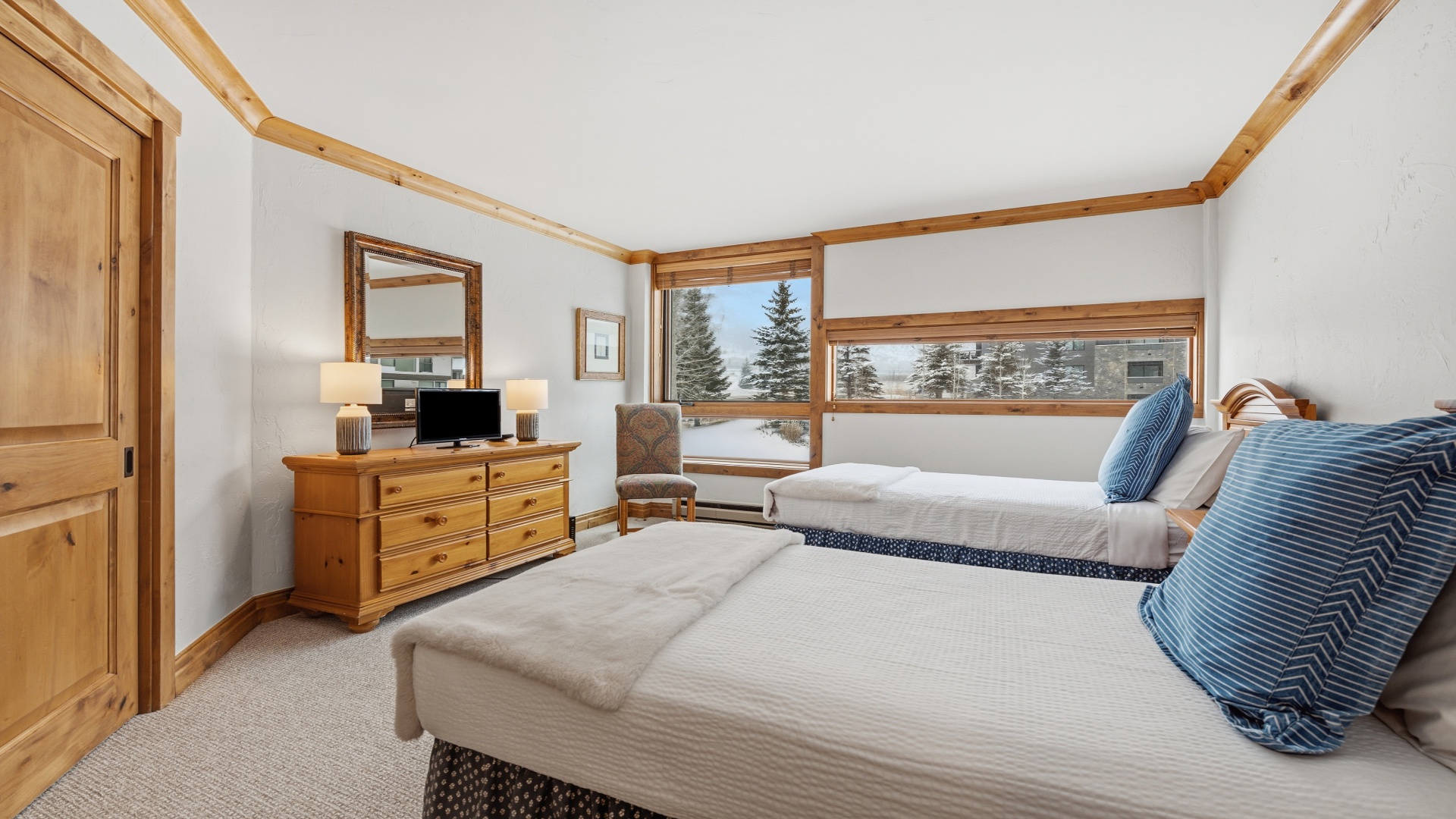 Unwind in your serene bedroom with stunning winter views. Two comfortable beds draped in soft linens invite restful sleep, while natural light fills the space through large windows overlooking snowy evergreens.