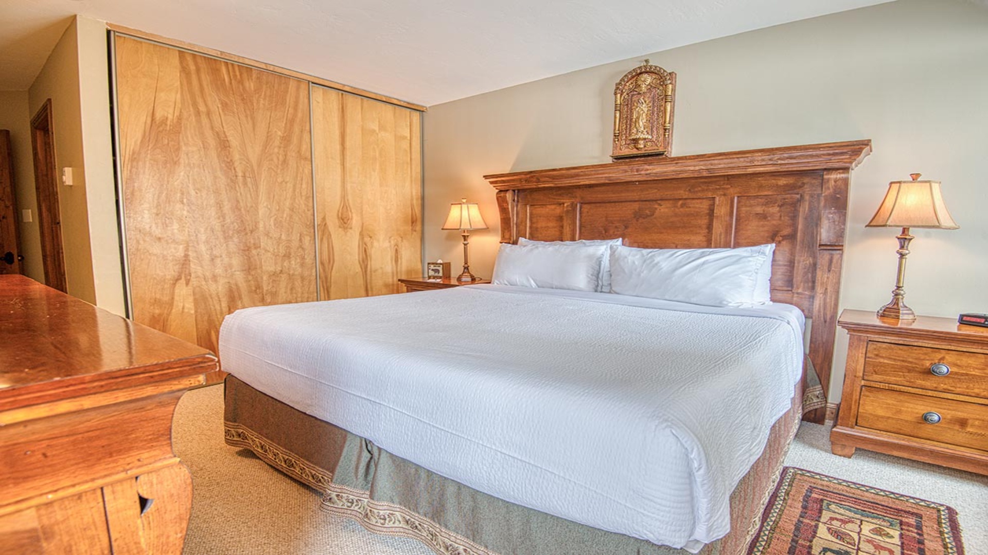 Your restful retreat features a plush bed with crisp white linens, warm wood furnishings, and soft bedside lighting that creates a cozy, welcoming ambiance.