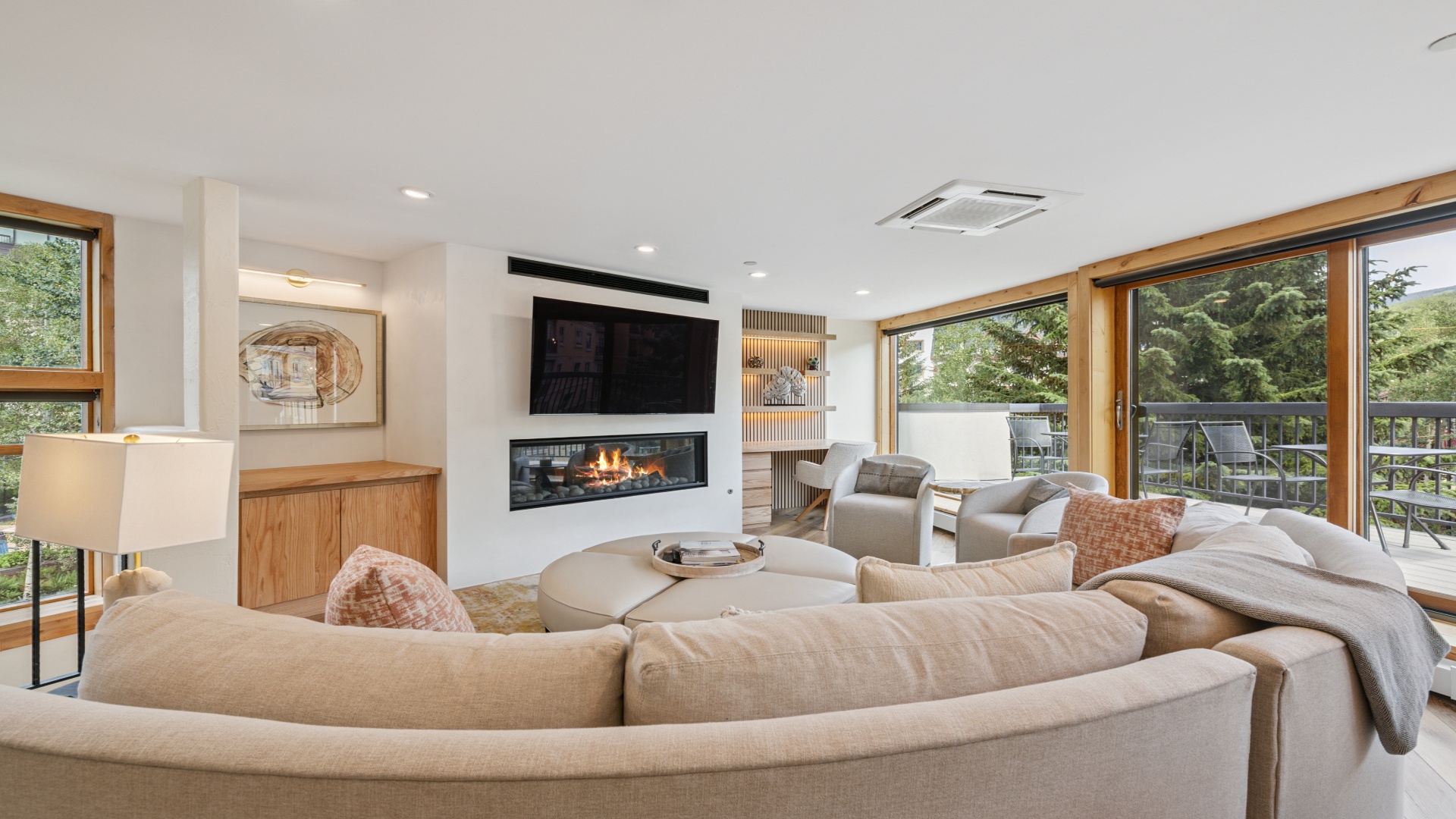 Unwind in your sunlit living room, where plush sofas curve around a warm fireplace and floor-to-ceiling windows frame lush greenery. Perfect for cozy evenings or bright mornings with coffee in hand.