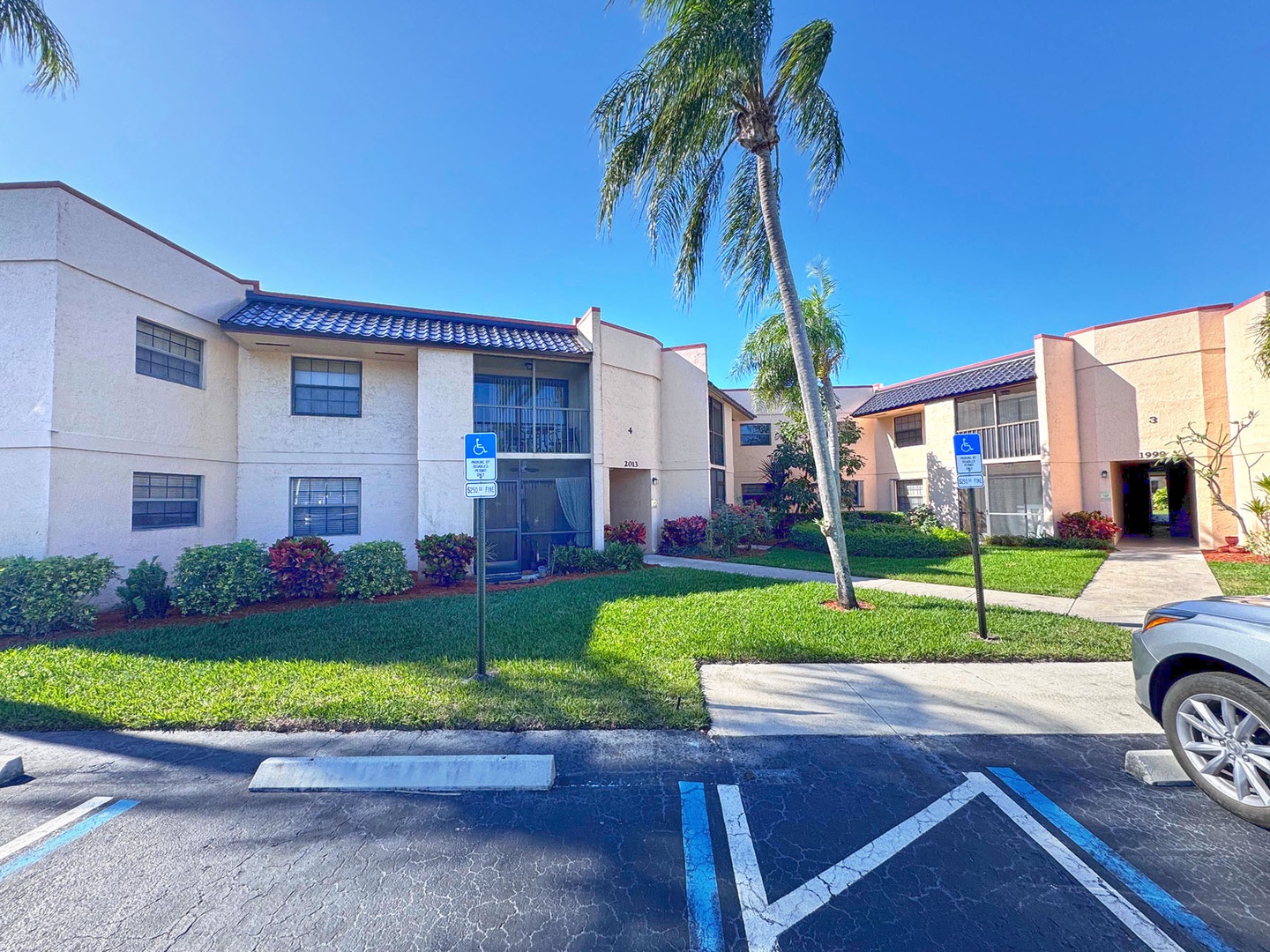 A sunny two-story building complex with tropical landscaping, accessible parking, and blue tile roofing in a residential neighborhood.