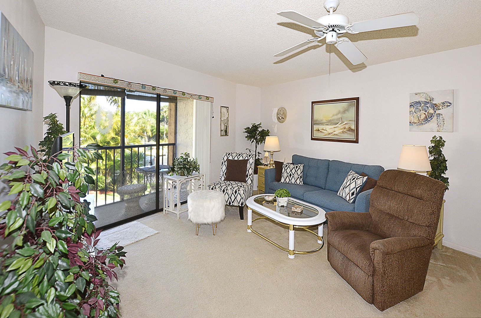 Relax in your bright living room with comfortable seating, ceiling fan, and serene balcony views of tropical greenery.
