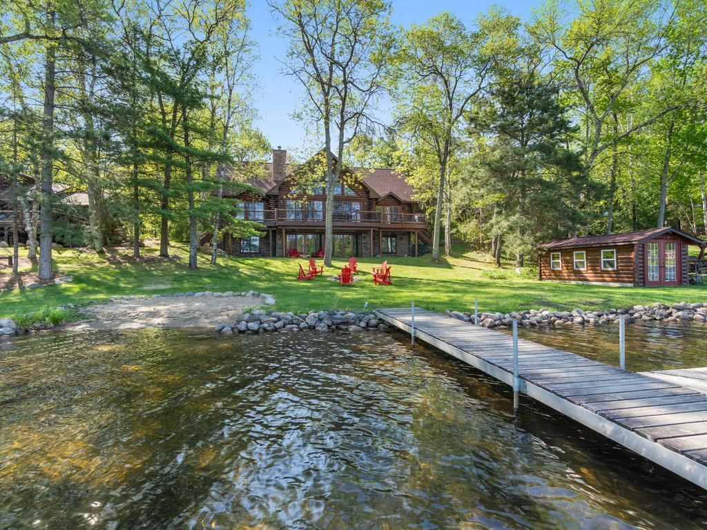 A peaceful lakefront property surrounded by towering trees, featuring a private dock and charming log cabin architecture nestled among lush greenery.