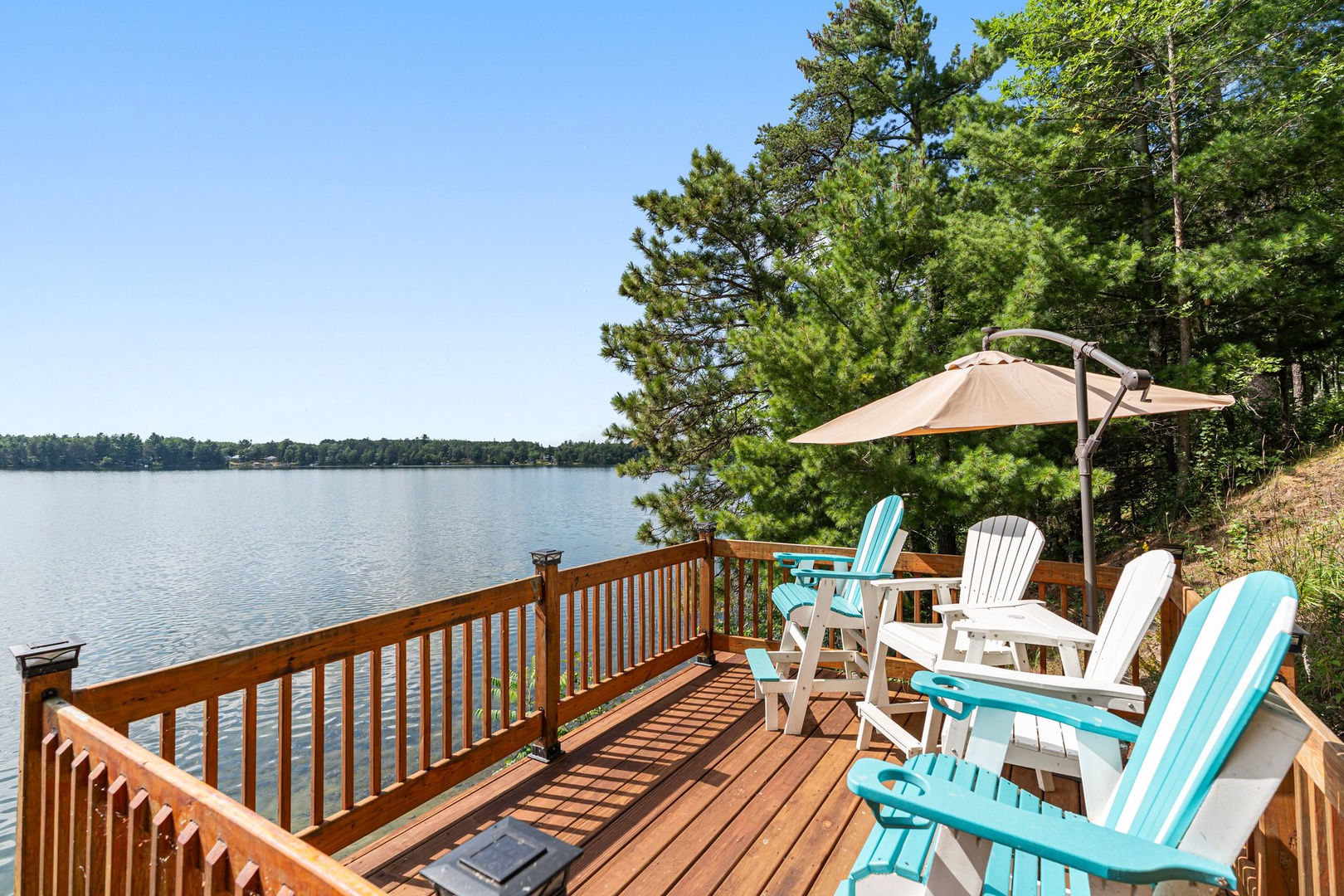Relax on your private lakefront deck with colorful Adirondack chairs and umbrella shade, watching peaceful waters through towering pines.