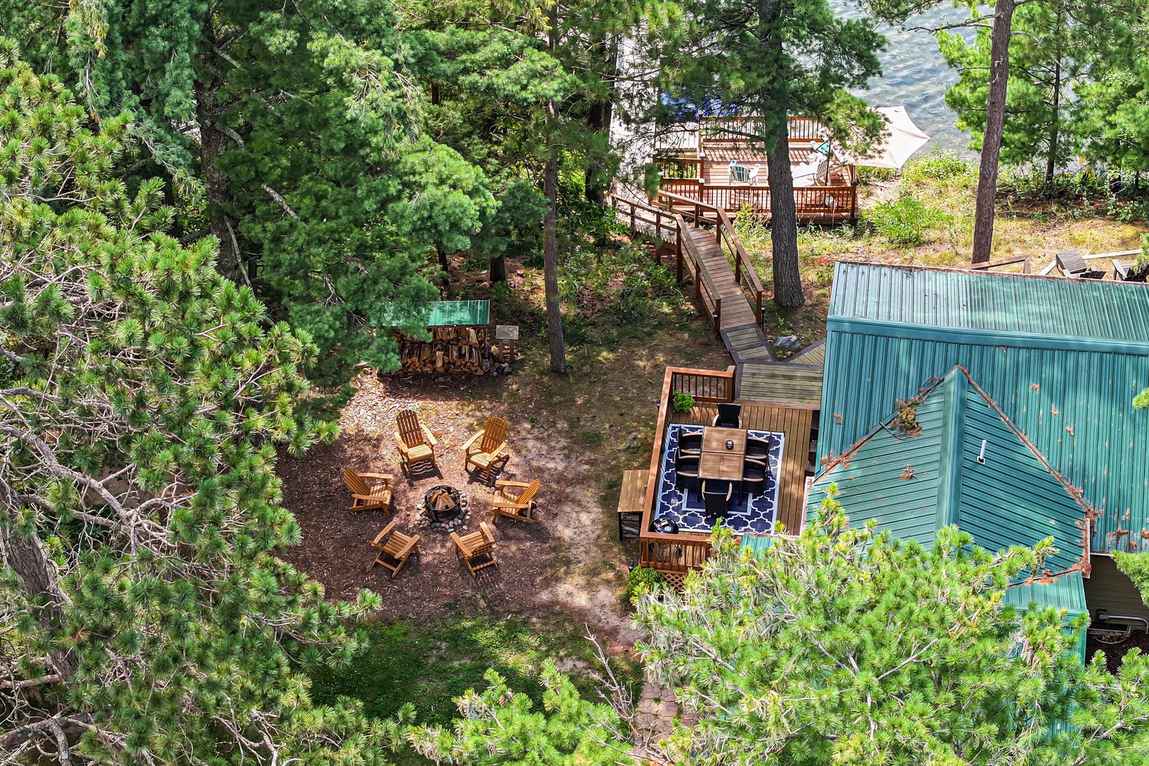 Aerial view of lakefront cabin property featuring outdoor fire pit seating area, hot tub deck, and private waterfront access through the forest.
