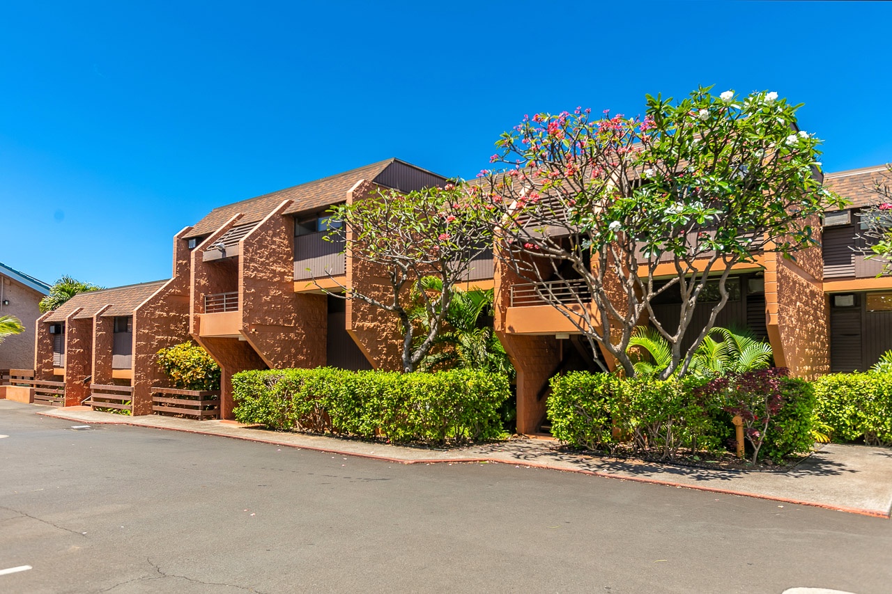 The property features warm brick architecture surrounded by vibrant tropical landscaping and manicured hedges under sunny skies.