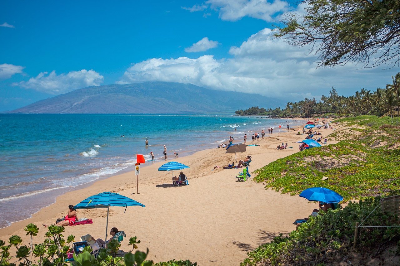 Sandy beach stretches along turquoise waters with mountain backdrop and colorful umbrellas dotting the shoreline.