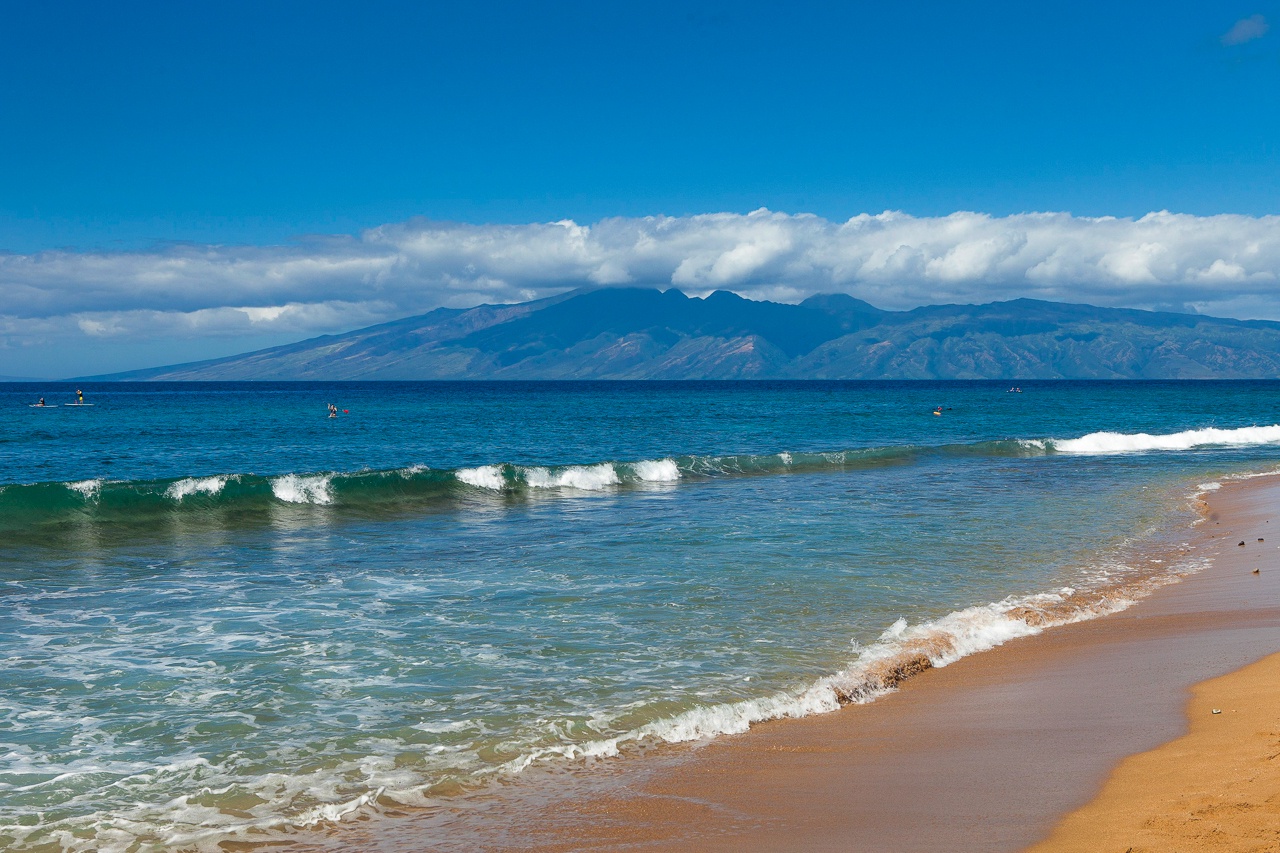 Pristine beach shoreline with turquoise waters meeting soft golden sands, surrounded by majestic mountain scenery and endless blue skies.