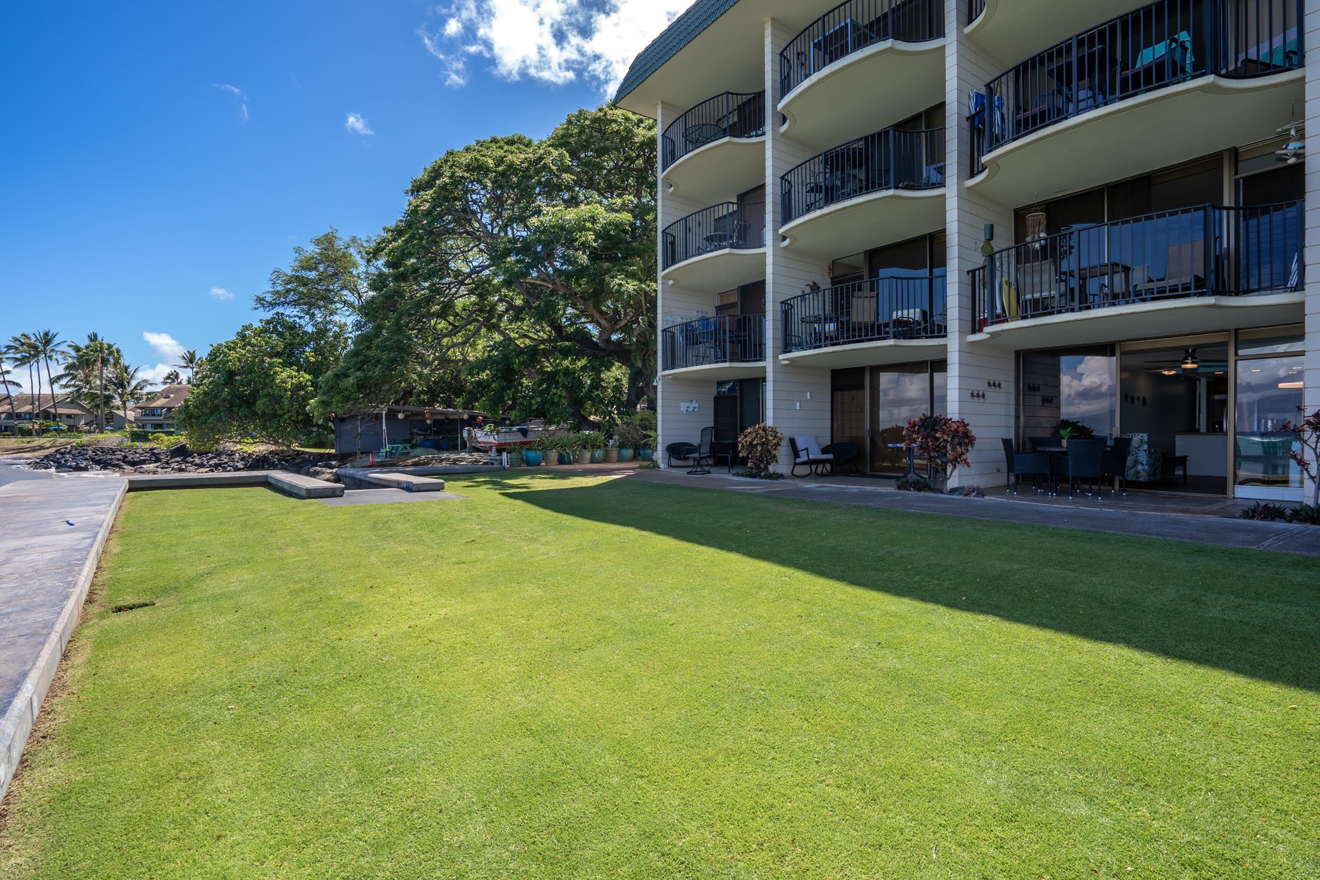 Multi-story vacation condo building with private ground-floor patios opening onto a spacious lawn, framed by mature trees and tropical landscaping under a clear Hawaiian sky.