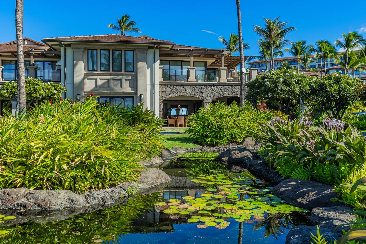 Beachfront villa surrounded by lush tropical gardens. Palm trees frame the building against clear blue skies and ocean views beyond.
