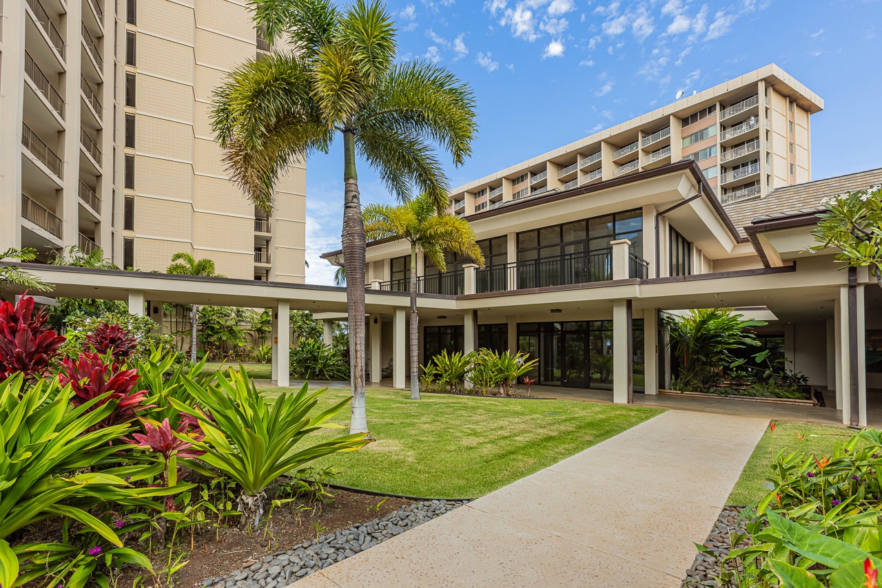A sun-soaked resort complex surrounded by manicured lawns and vibrant tropical gardens, with swaying palms and colorful foliage framing the modern architecture beneath a brilliant blue sky.