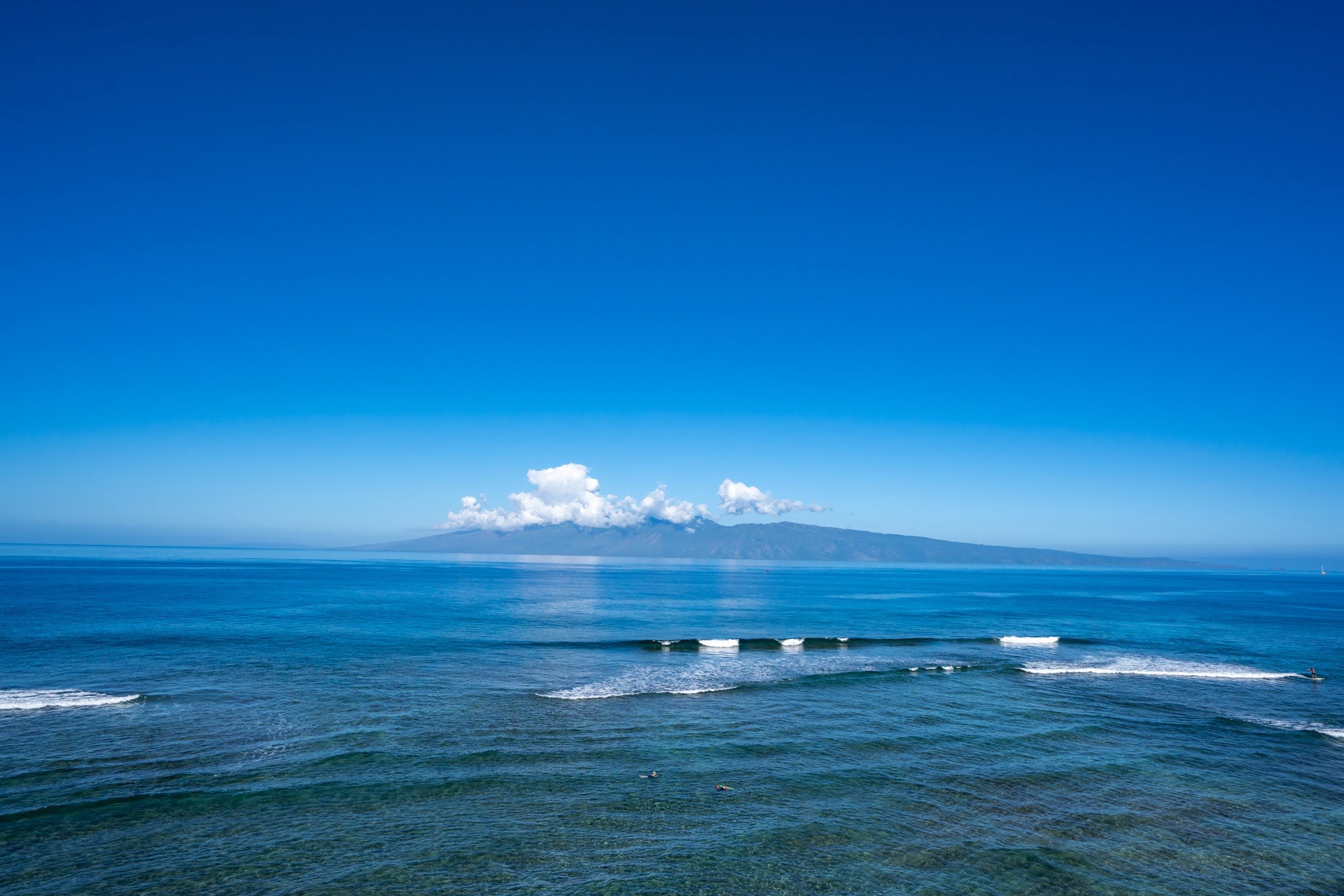 A stunning panoramic scene of the Pacific Ocean unfolds with gentle surf rolling toward shore and a distant island rising beneath soft clouds.