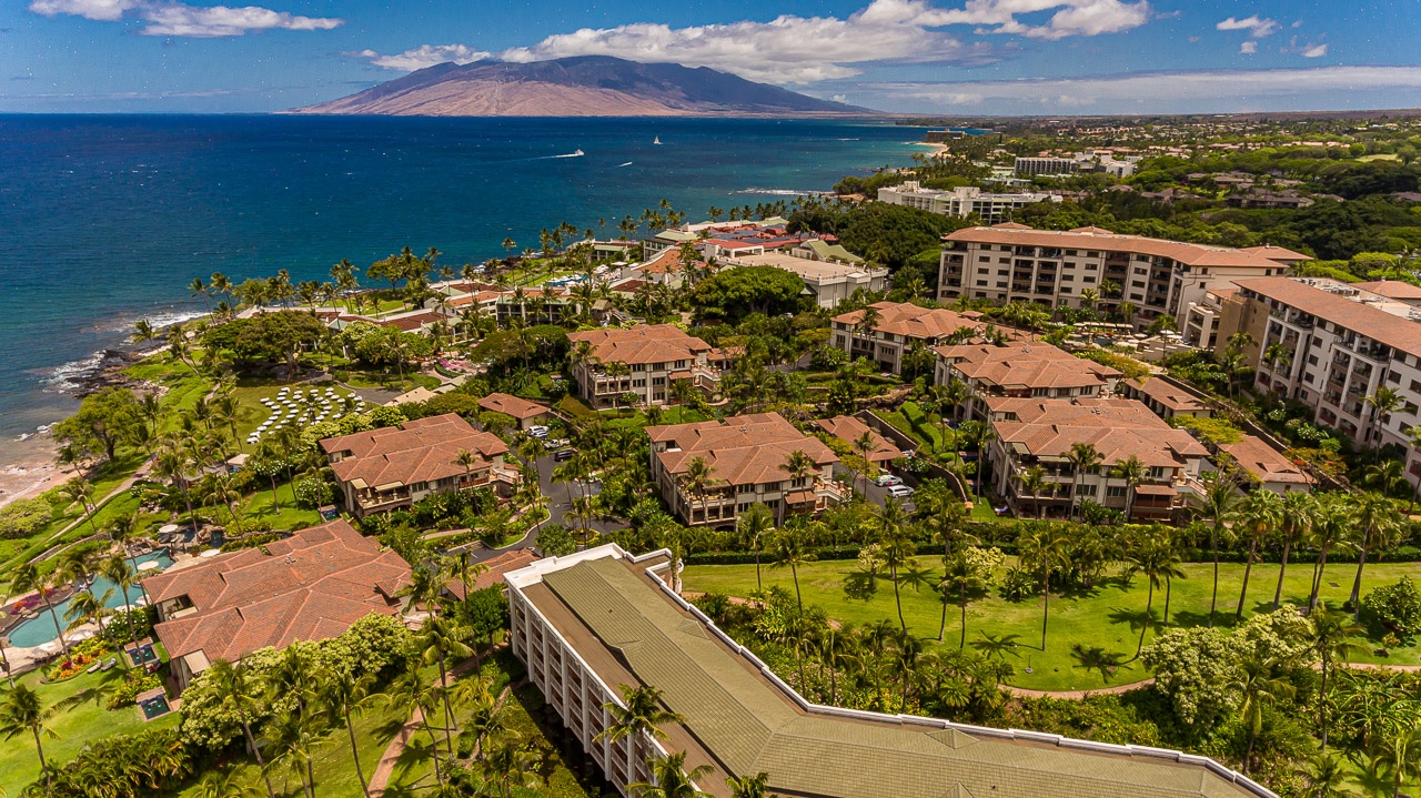 Aerial view of a Maui resort community stretching along the coastline, with lush tropical gardens, red-tiled roofs, and the island of Lanai visible across the sparkling Pacific waters.