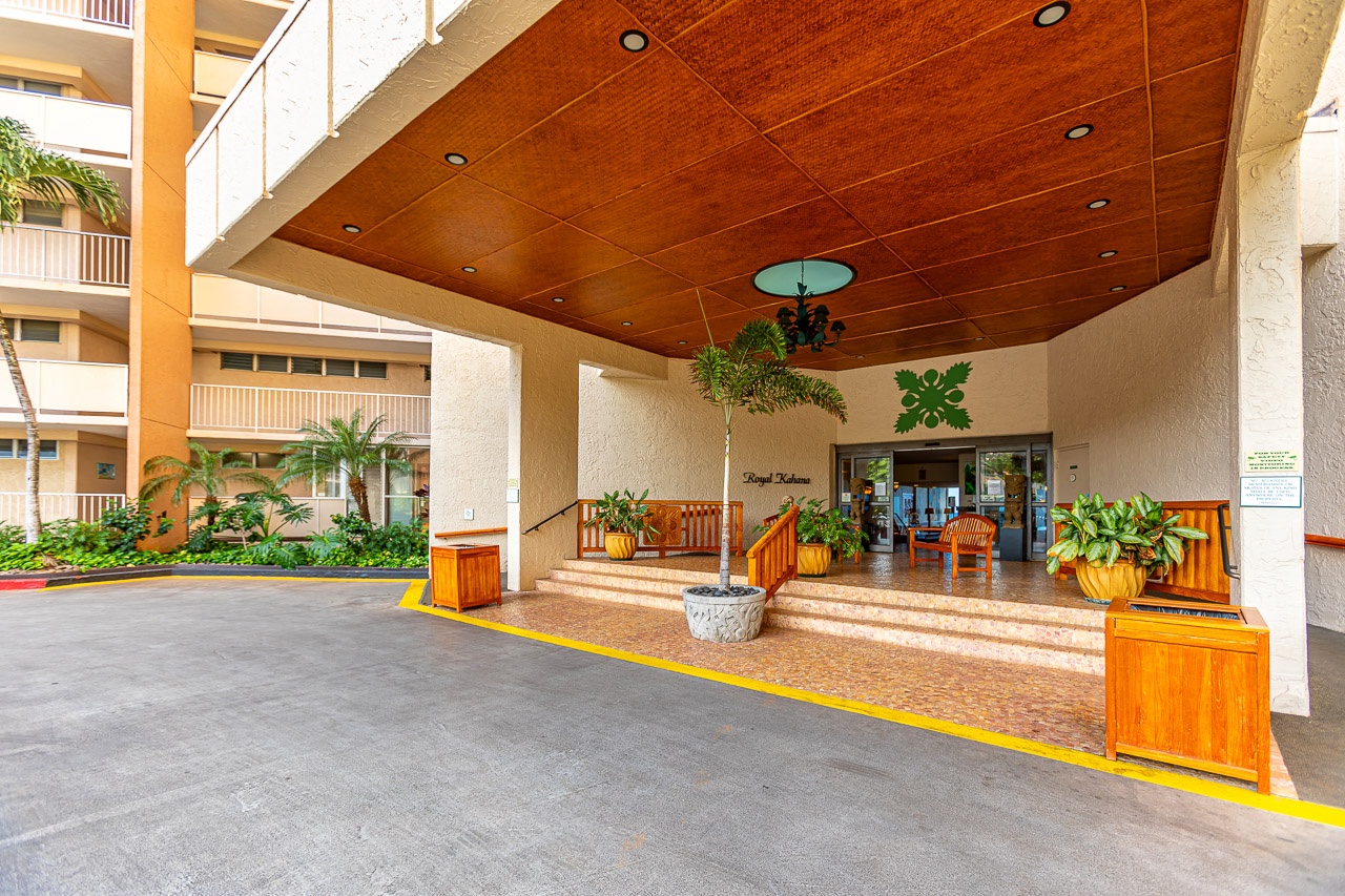 A covered entrance with warm tropical accents provides a welcoming arrival area. Benches and lush potted plants frame the doorway.