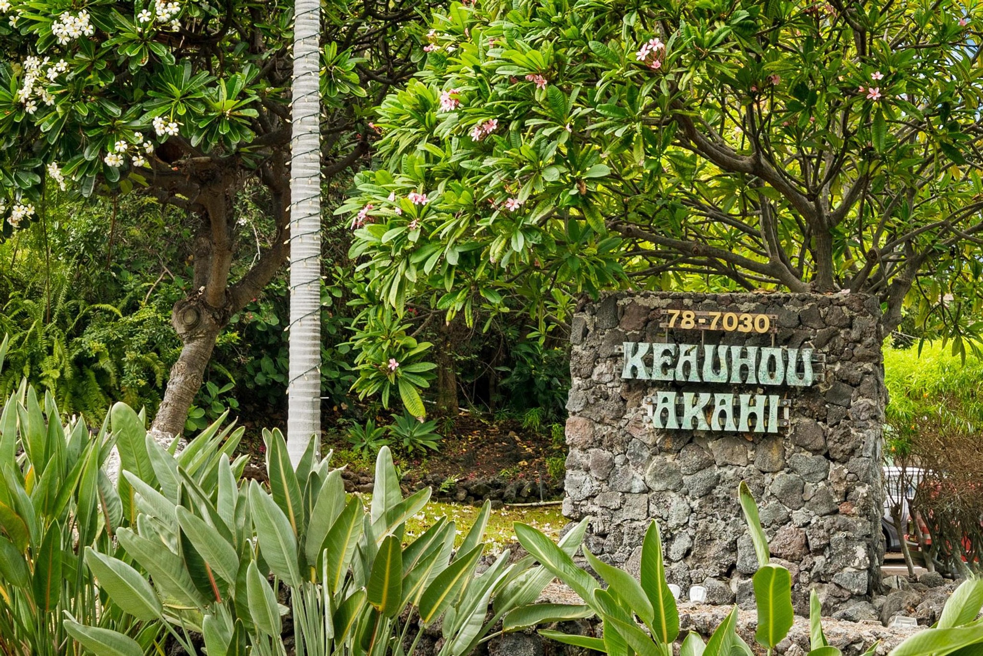 Stone entrance marker surrounded by lush tropical landscaping including flowering trees, palm, and bird of paradise plants.