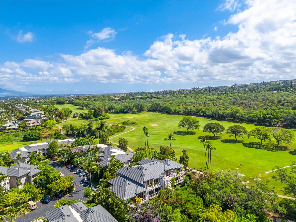 Aerial view of the property nestled within a lush residential area adjacent to a championship golf course, surrounded by tropical greenery and mountain vistas under open Hawaiian skies.