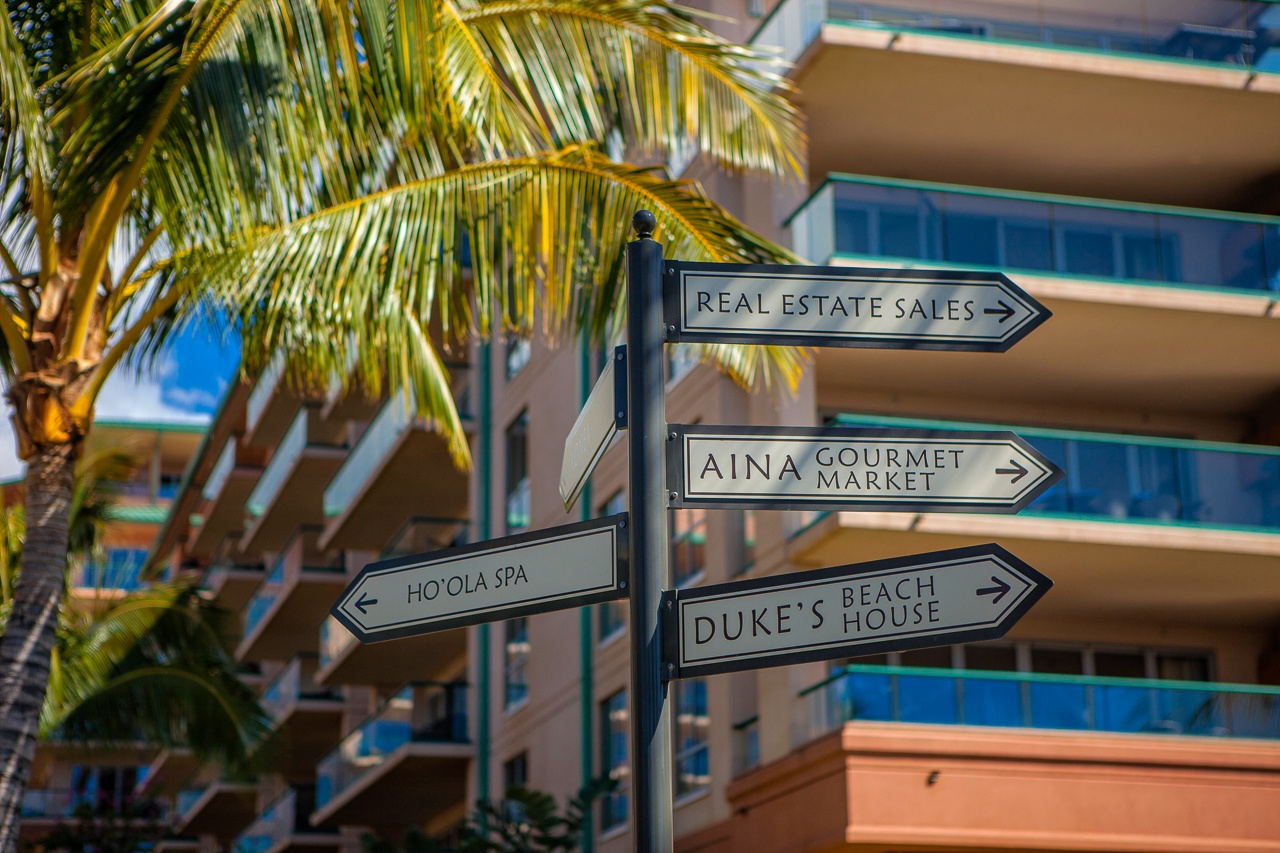 Directional signage near the property entrance guides visitors to local attractions including the Aina Gourmet Market, Ho'ola Spa, and Duke's Beach House, with palm trees framing the scene.