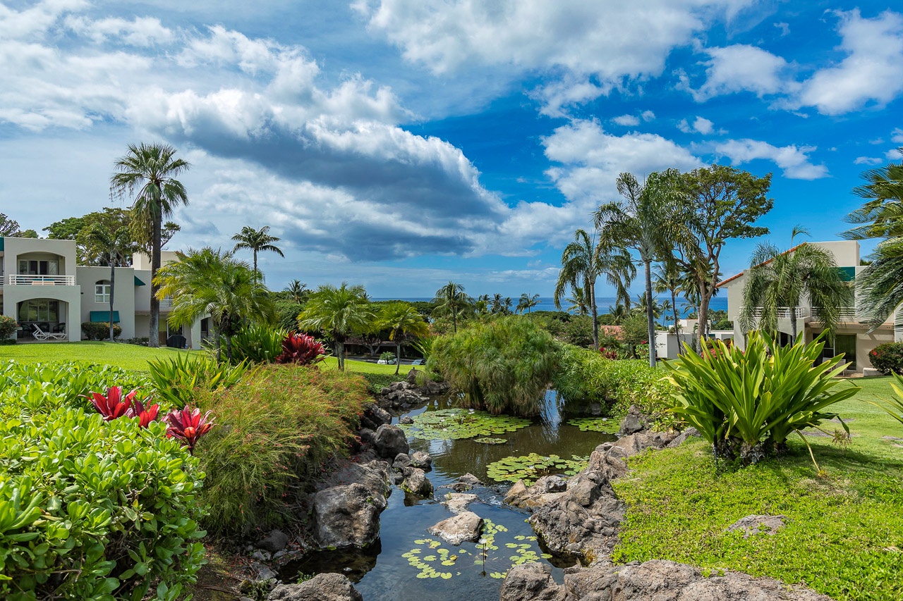 A serene waterway winds through lush tropical gardens, framed by palm trees and colorful blooms, with the sparkling ocean visible on the horizon beyond manicured lawns.