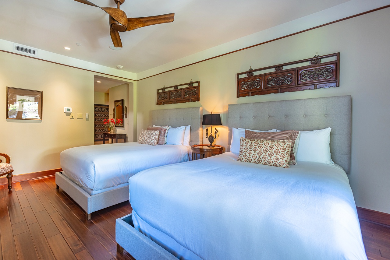 Your spacious bedroom features two plush beds with elegant carved wood accents overhead, warm hardwood floors, and gentle ceiling fan breezes to lull you to sleep after a day exploring paradise.