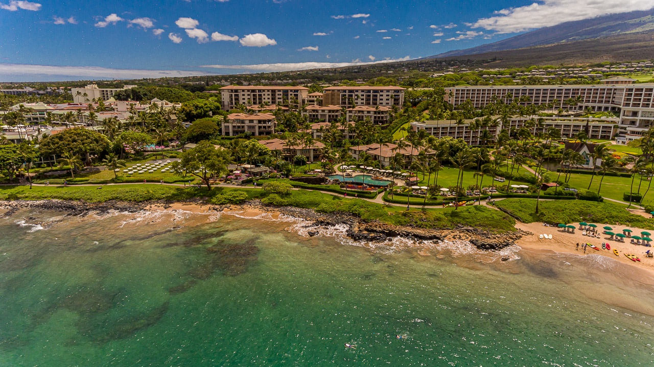 A beachfront resort complex nestled between turquoise waters and lush tropical grounds, with mountain views stretching across the horizon.