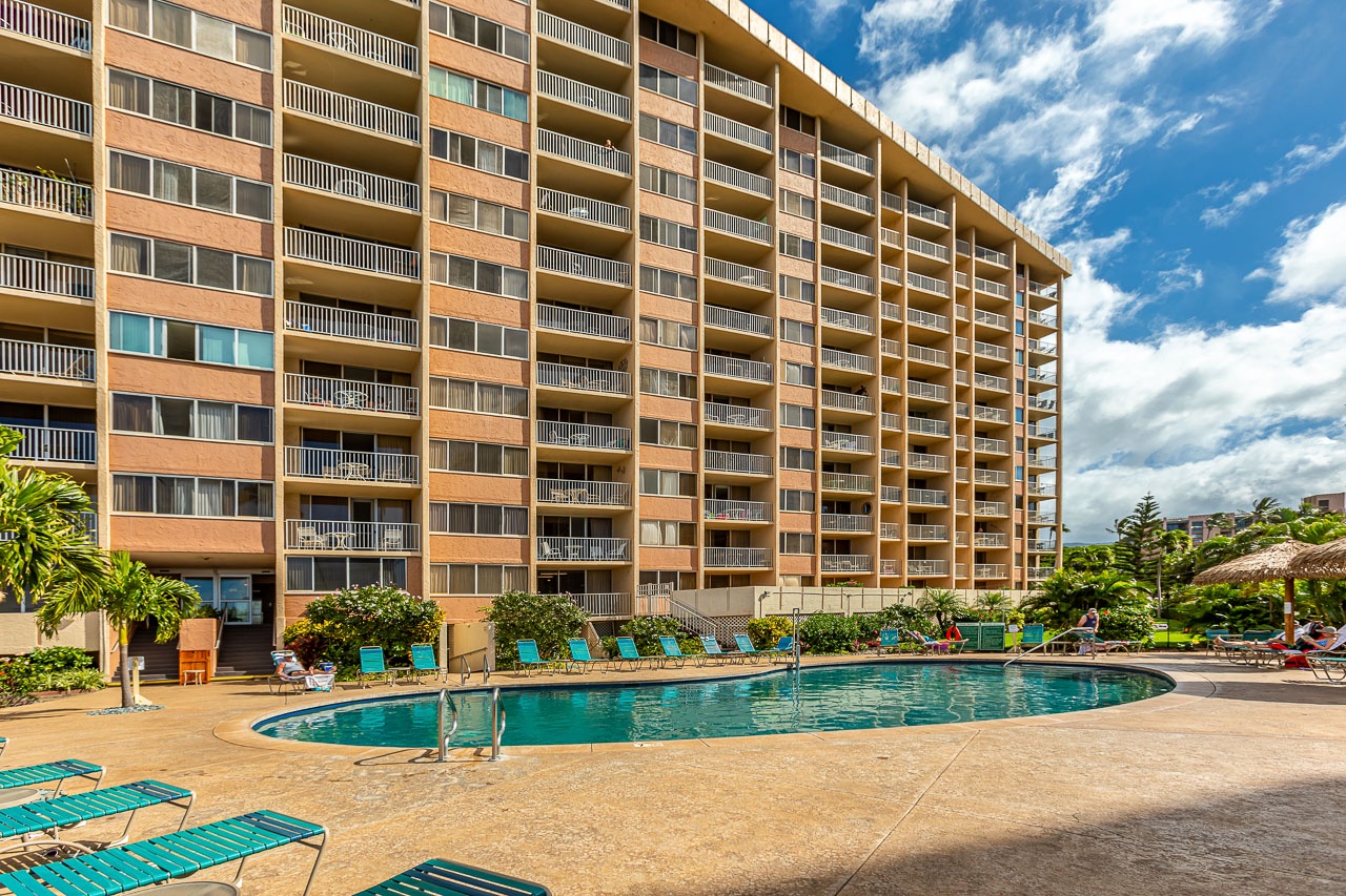 The property features a spacious outdoor pool surrounded by tropical landscaping and loungers, set against a multi-story building with private balconies.