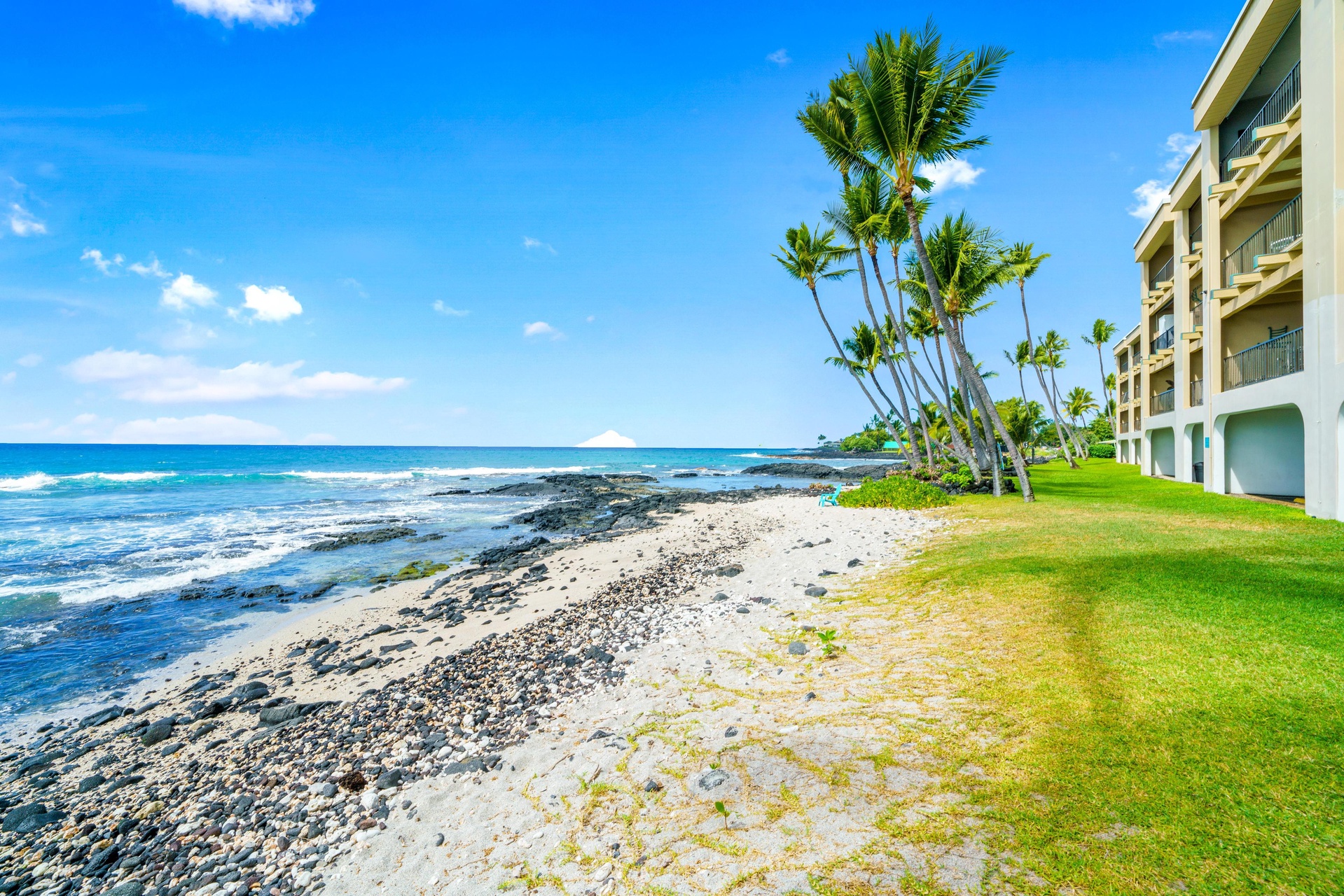 This oceanfront property offers direct access to a tranquil pebbled shore where turquoise waves meet tropical palms. The building sits on manicured lawns just steps from the sea.
