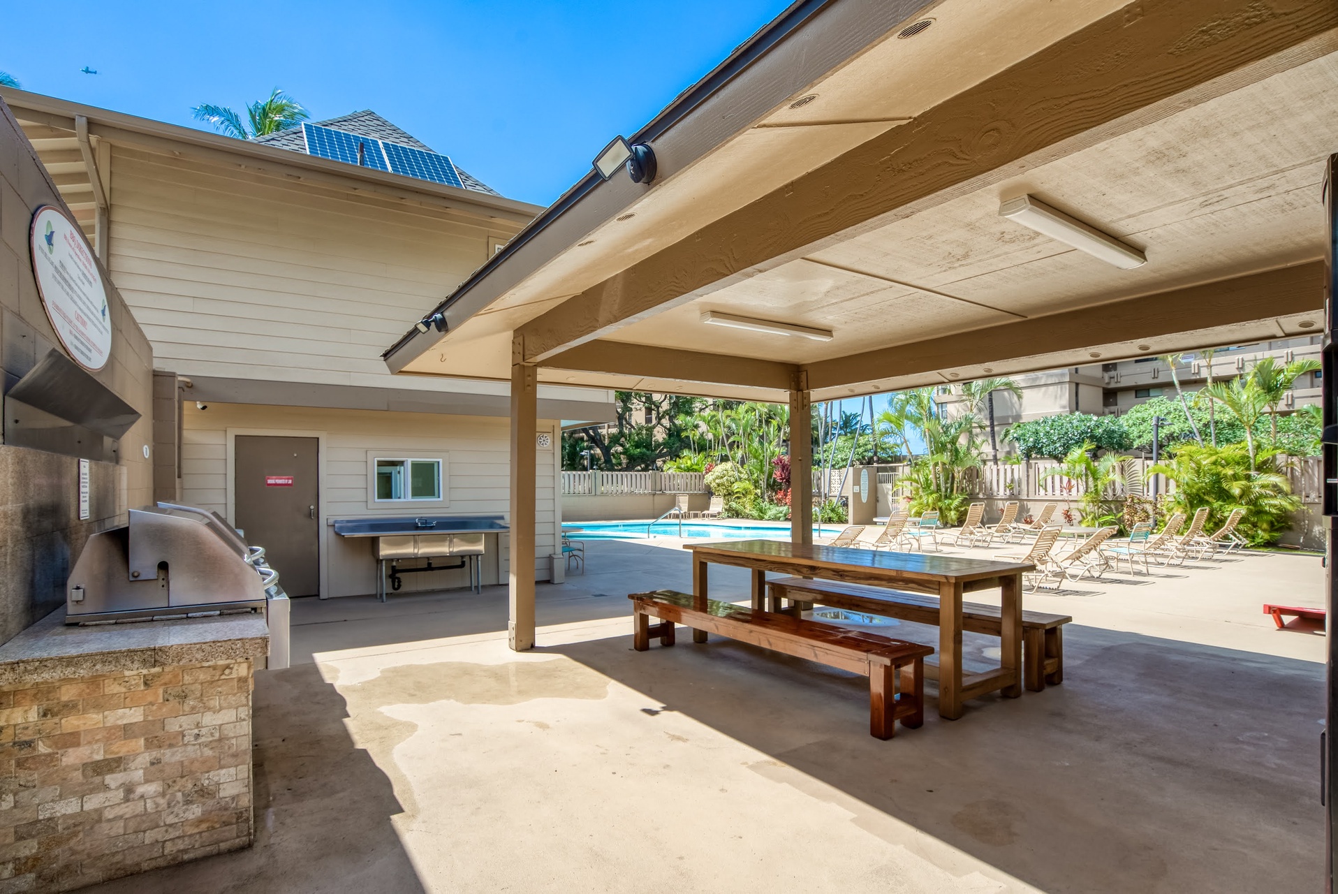 Step under the covered pavilion to find a rustic dining table and BBQ grill, with the sparkling pool just beyond and tropical palms swaying in the breeze.
