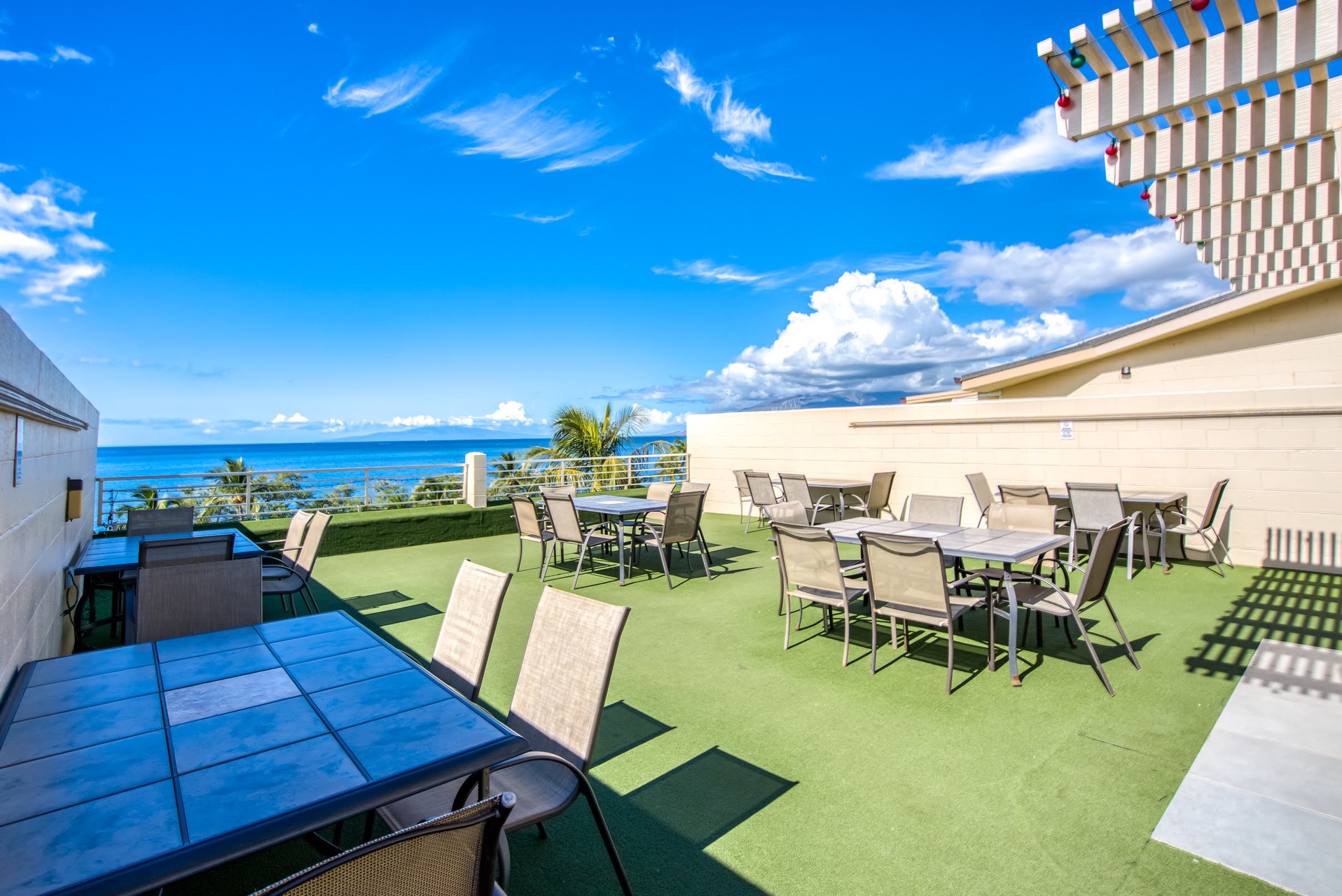 Rooftop terrace overlooking the ocean with dining tables, lounge seating, and striped awning providing a breezy outdoor gathering space.