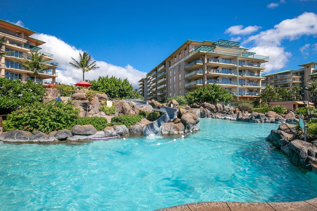 Tropical resort featuring a winding pool with waterslide, lush landscaping with palm trees, and modern multi-story buildings offering balcony accommodations under sunny skies.
