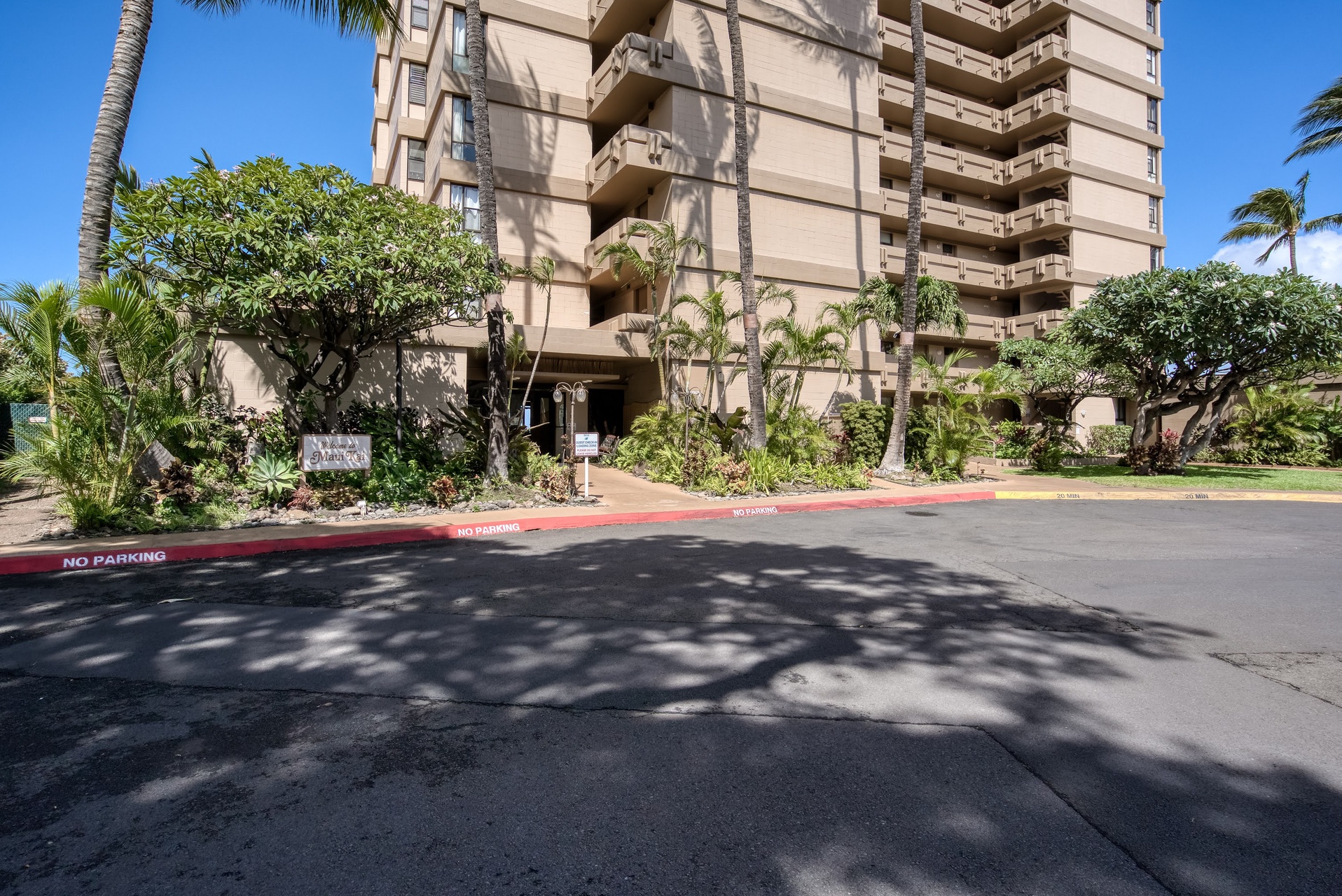 The property building entrance is surrounded by lush tropical landscaping, with palm trees and greenery framing the façade in a sunny Hawaiian setting.