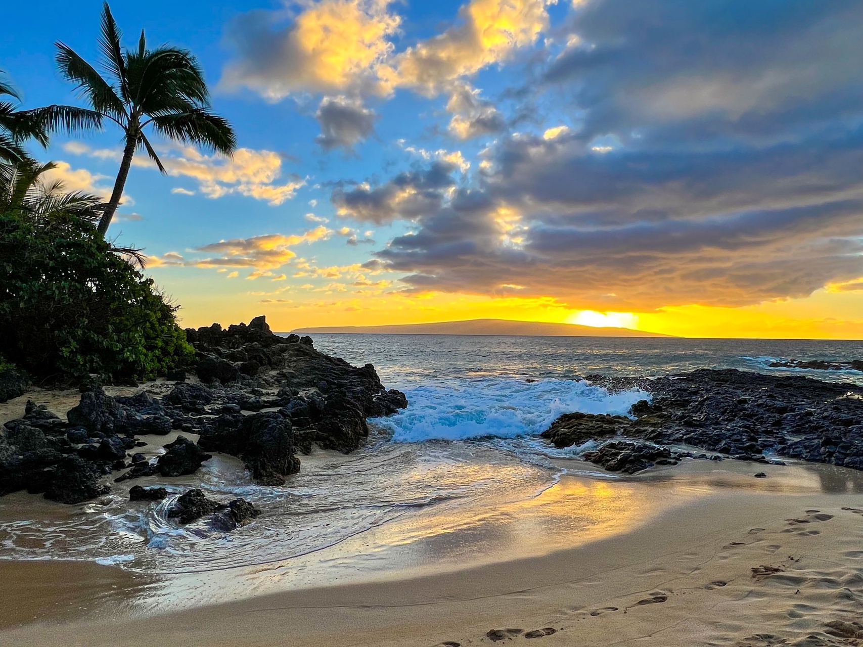 Golden sunset illuminates the secluded beach cove, where lava rock formations frame soft sands and swaying palms mark the tropical shoreline nearby.