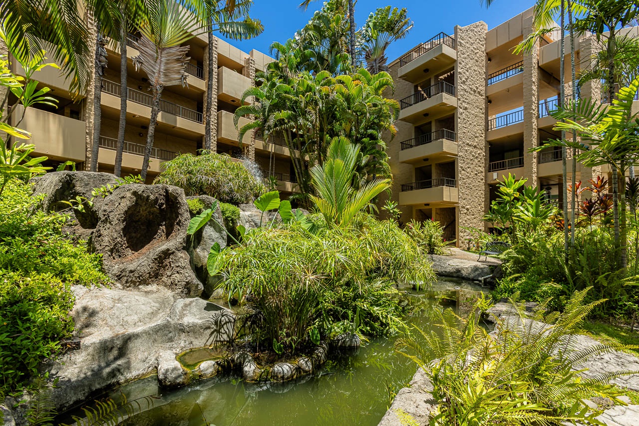 A serene inner courtyard features lush tropical landscaping with palm trees, ferns, and vibrant greenery surrounding a tranquil pond with natural rock formations.