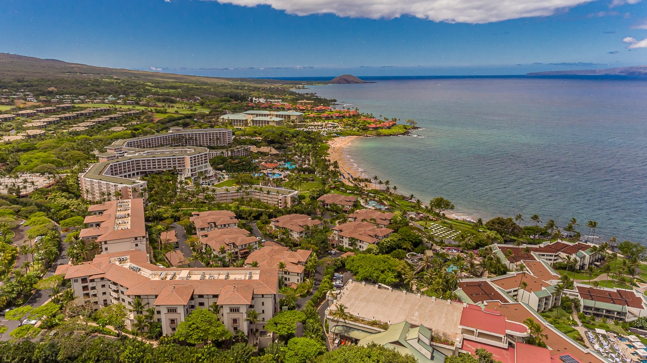 Expansive coastal resort area with lush tropical grounds nestled between verdant hillsides and tranquil ocean waters, featuring a pristine beachfront and island views on the horizon.